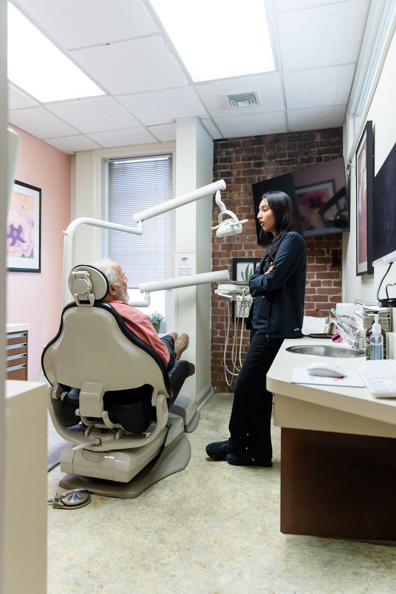 Dentist office with patient in chair and dentist standing nearby under bright ceiling lights.