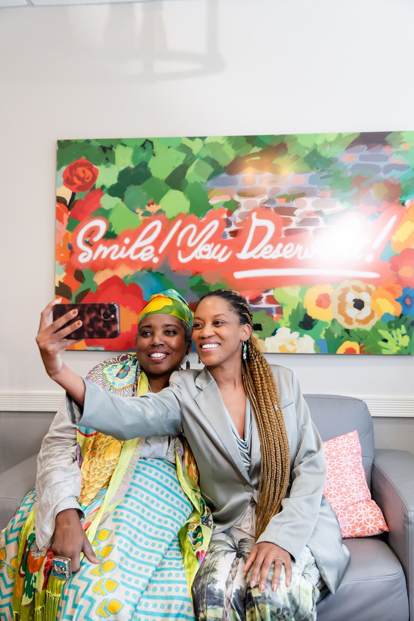 Two smiling people seated on a couch in front of a colorful floral “Smile Now, Dance...” sign.