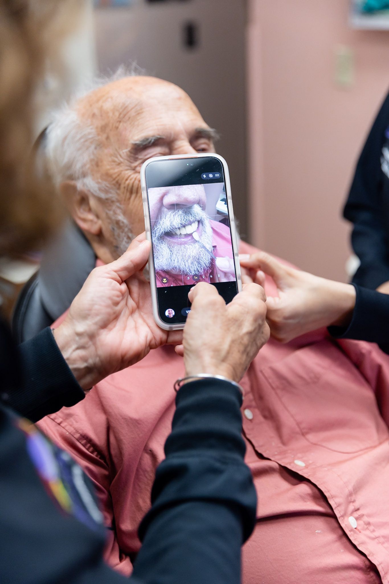 Person photographing an elderly man in a pink shirt with a smartphone indoors