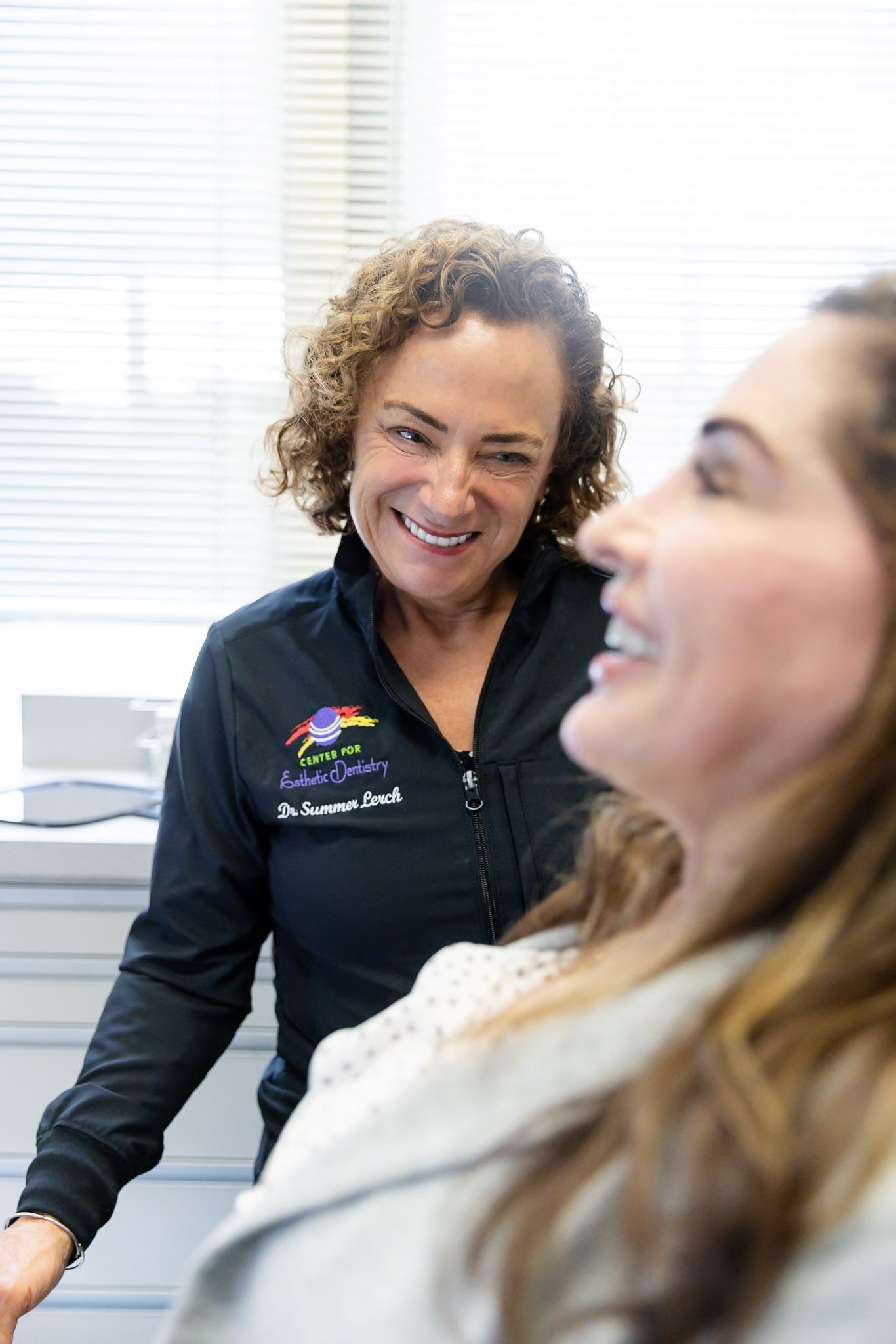 Two women smiling in a bright office, one in a black jacket