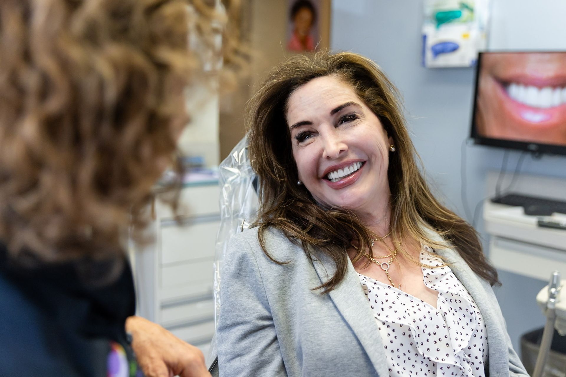 Woman smiling in a dental office, seated while speaking with someone nearby.