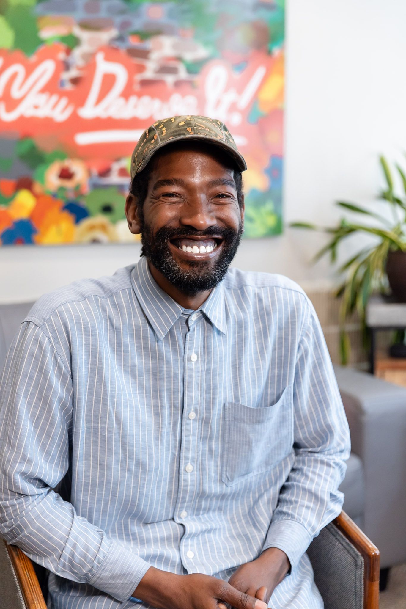 Smiling man seated indoors in a light blue checkered shirt and cap
