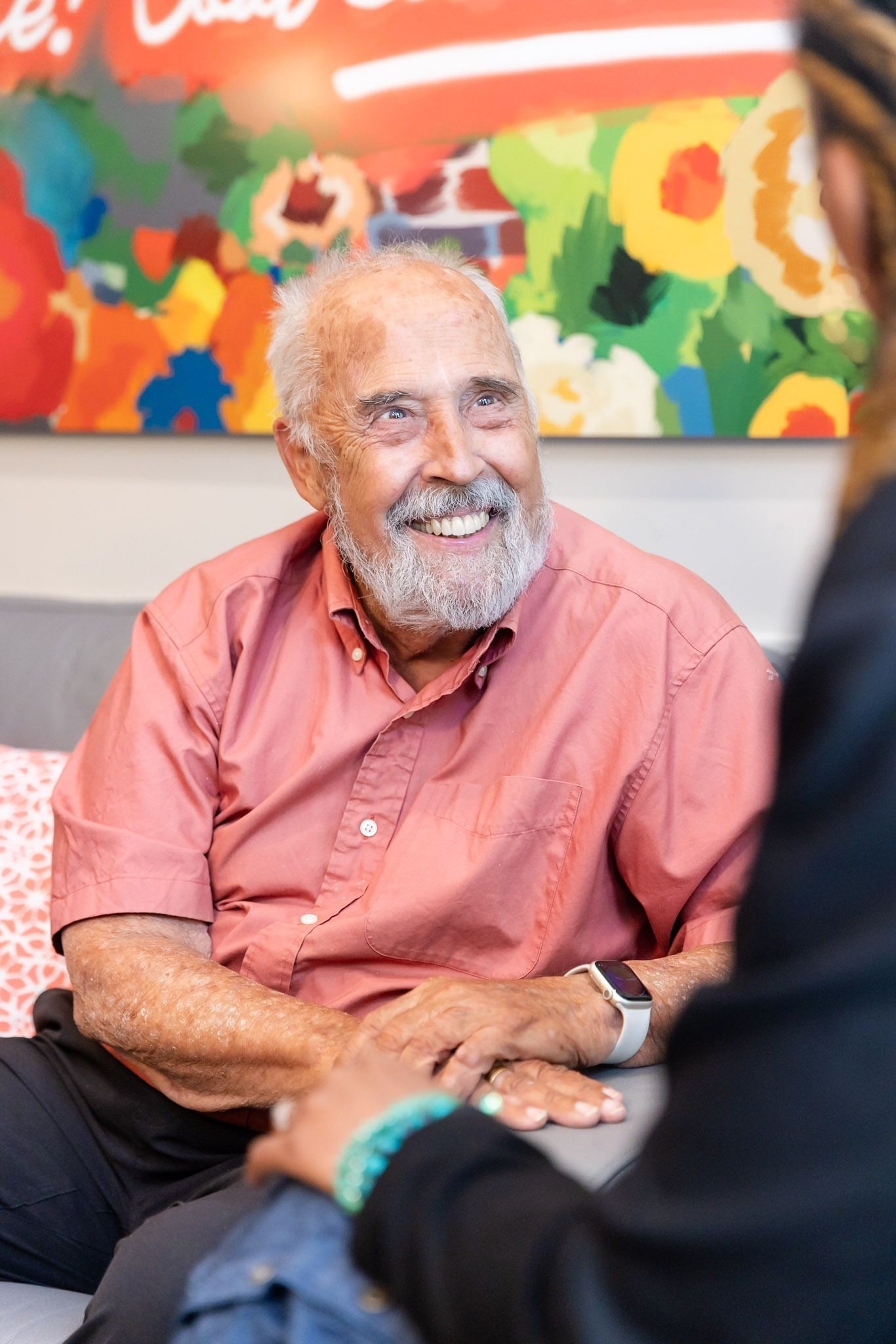 Smiling man in a pink shirt sitting indoors, with colorful abstract artwork behind him