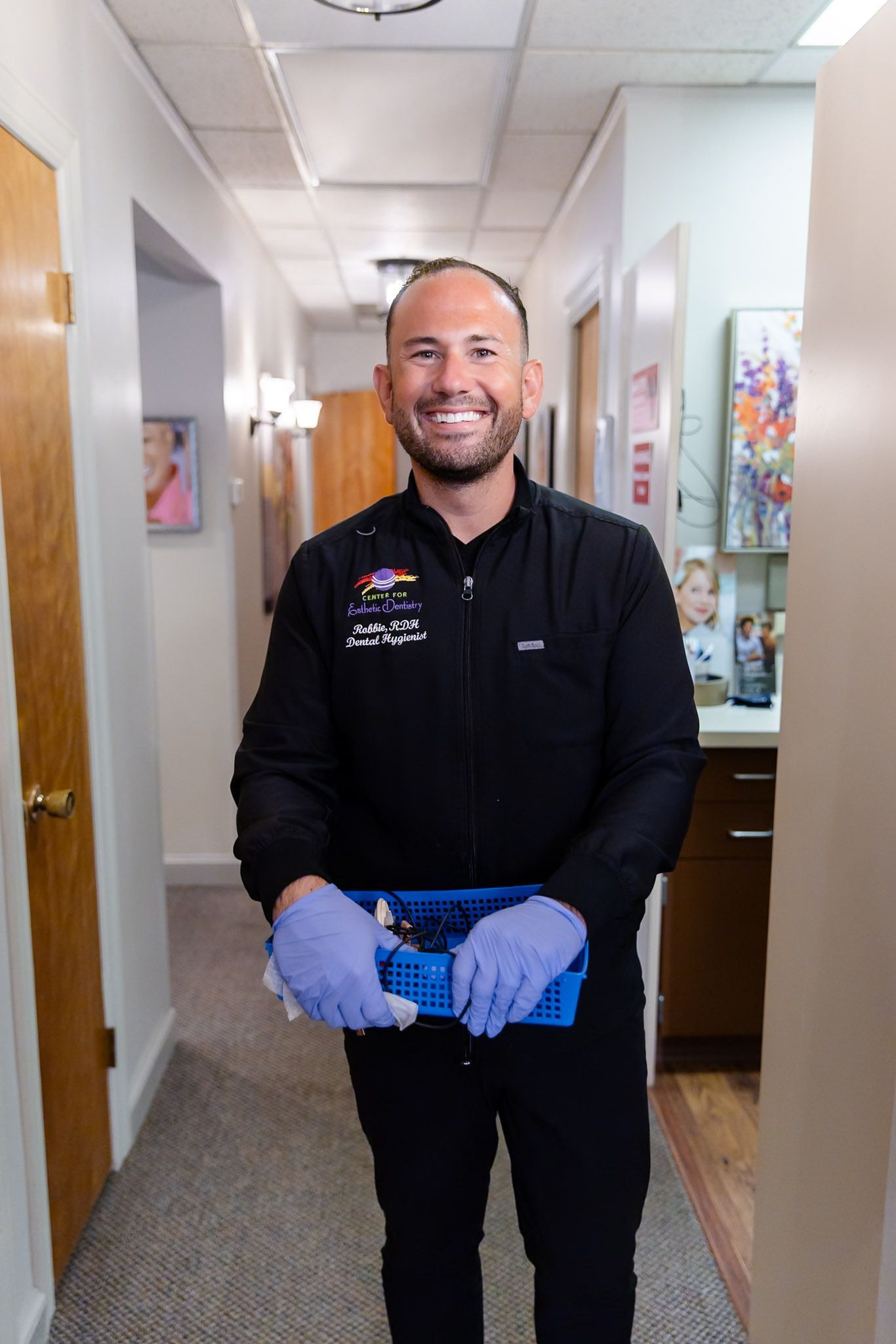 Smiling man in black uniform and blue gloves standing in a hallway holding a tray