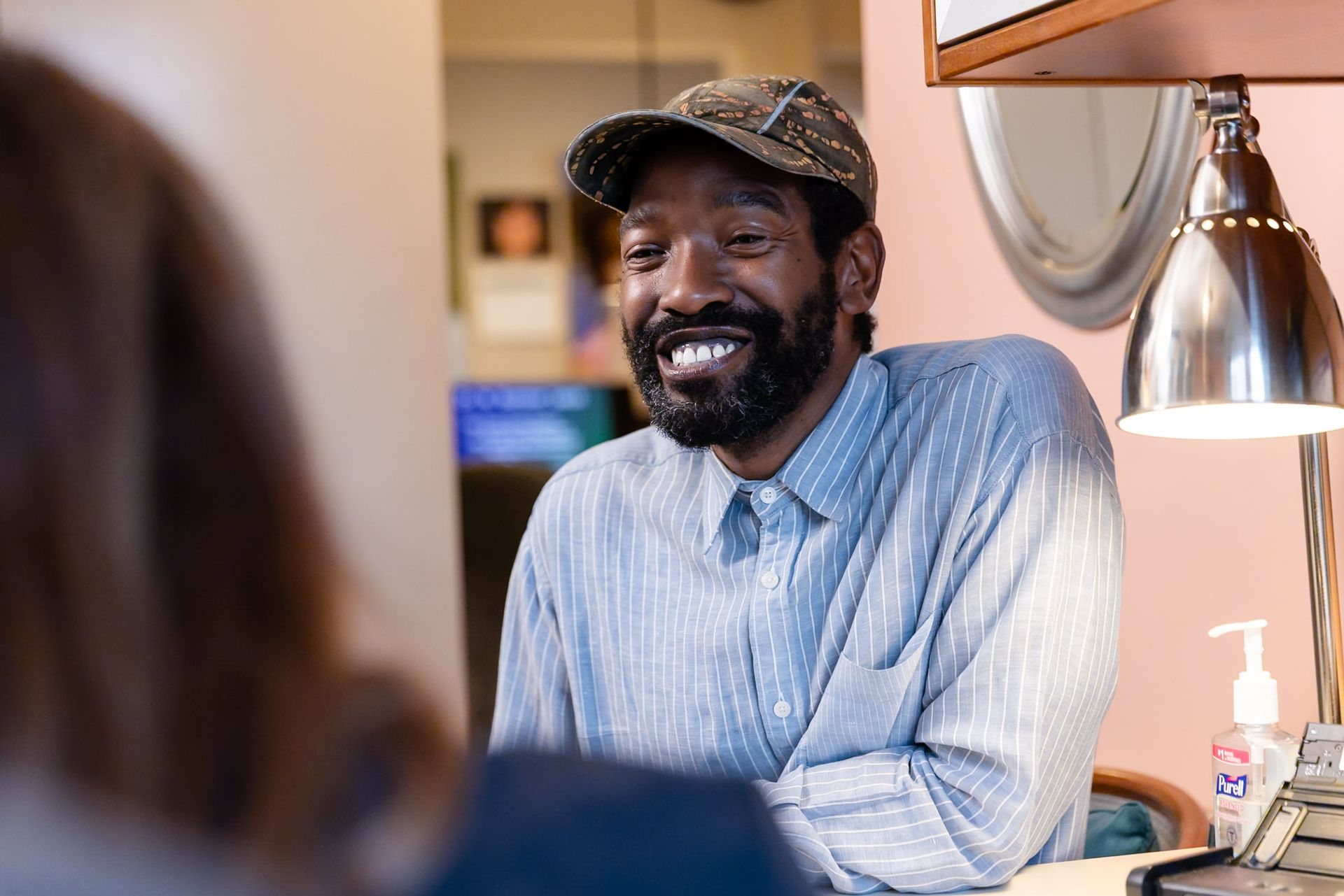 Smiling man in a cap sits at a table in a bright room, talking across from someone.