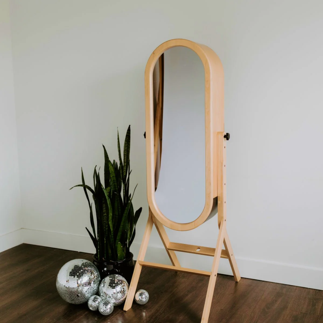 Wood-framed oval mirror on a stand beside a snake plant and silver ornaments on a wooden floor
