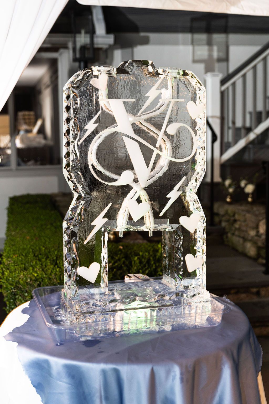 Decorative ice sculpture on a blue-draped table outdoors, with white swirls and heart shapes on a clear block