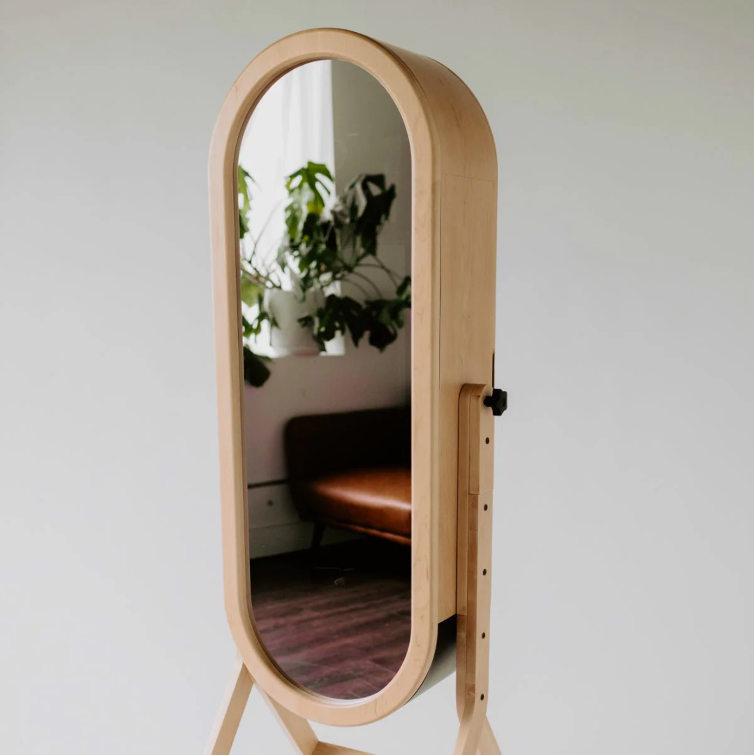 Tall wooden standing mirror with an arched frame reflecting a plant and sofa in a bright room