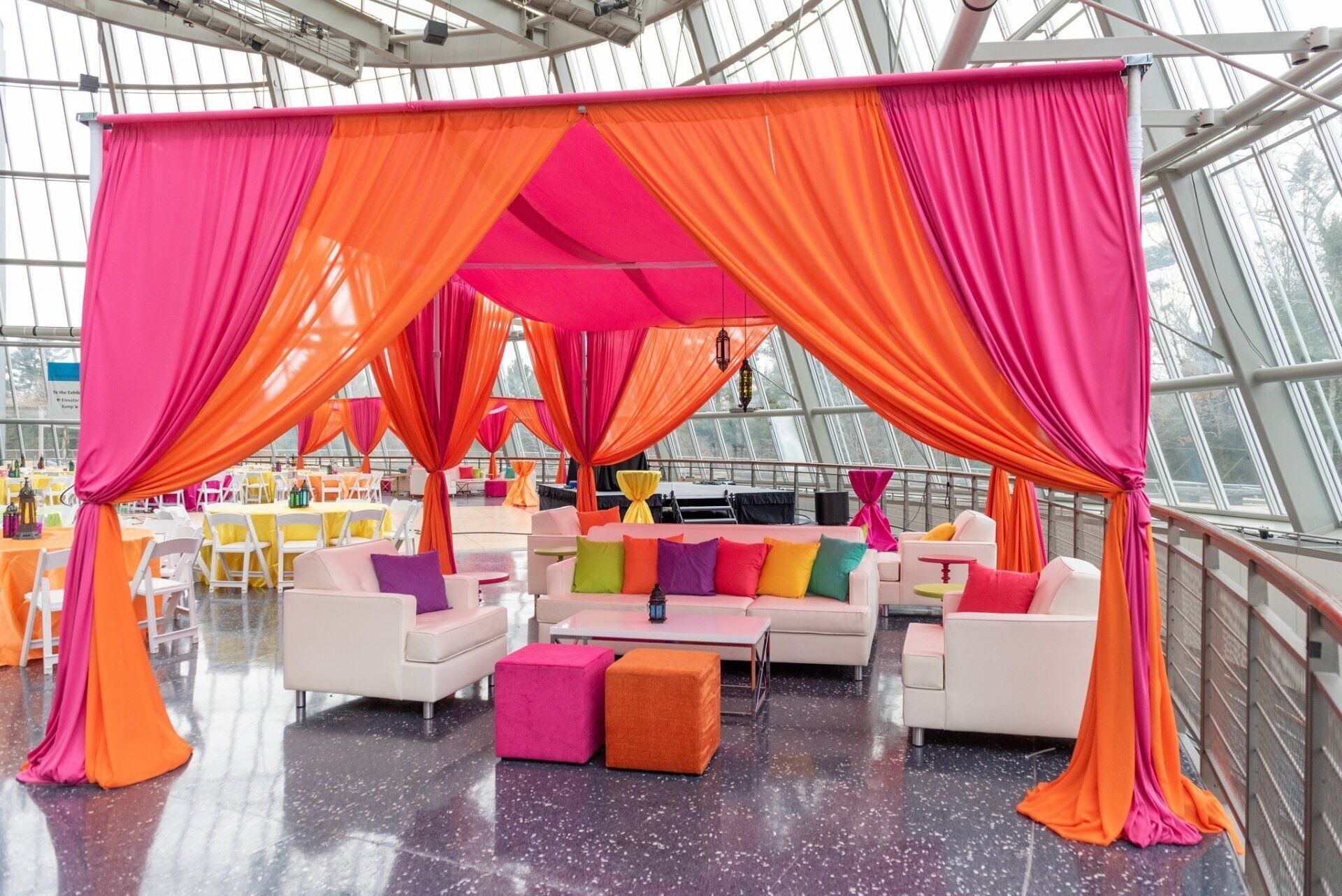 Colorful draped lounge with pink and orange fabric, white seating, and bright cushions in a glass atrium.