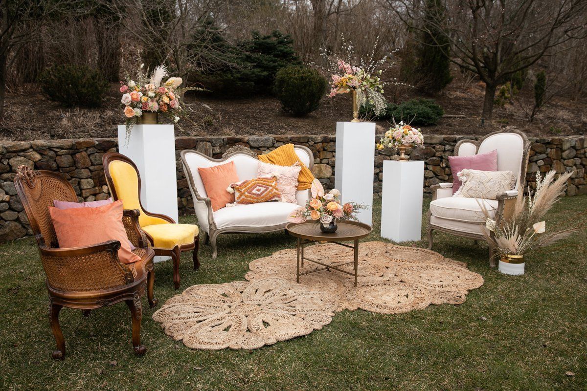 Outdoor floral lounge setup with white and peach chairs, tall flower vases, and woven rugs on grass