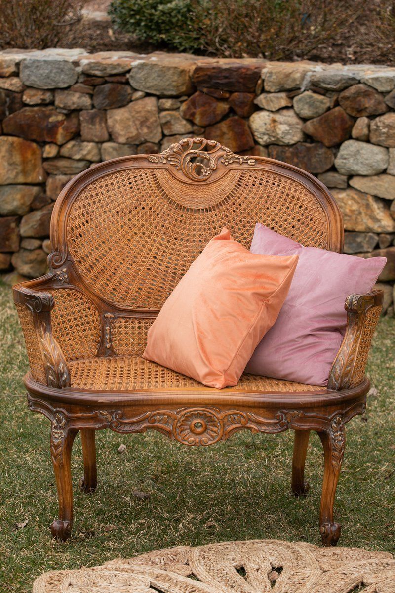 Wicker loveseat with peach and lavender pillows on grass before a stone wall