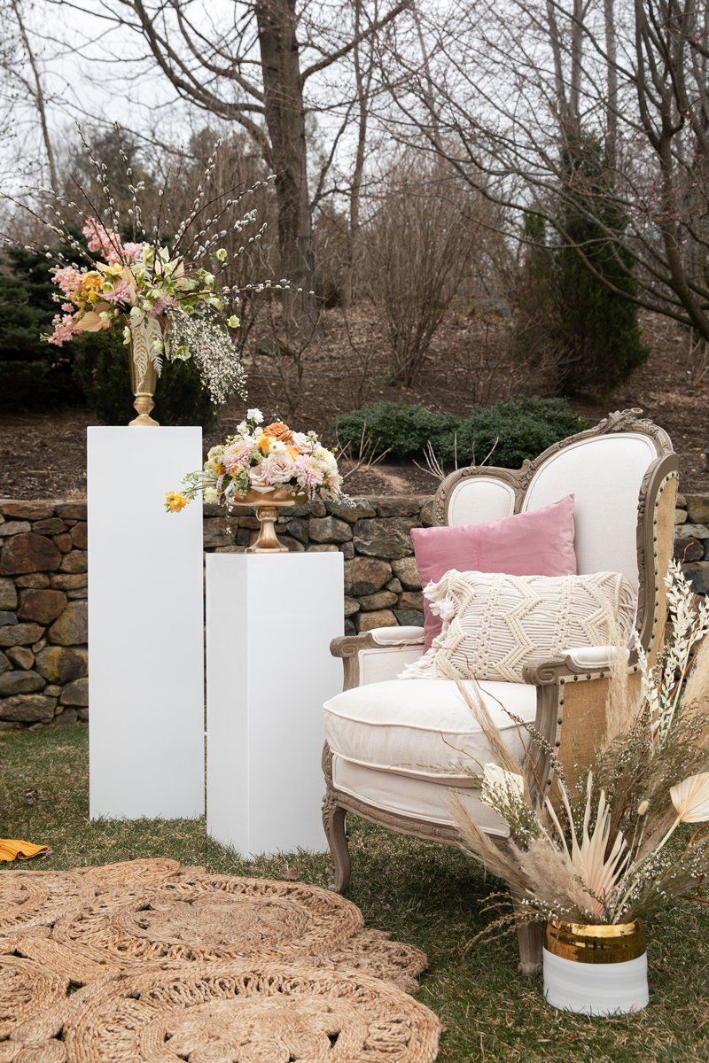 Decorated outdoor lounge with white chair, pink pillow, floral arrangements, and dried grasses in a garden setting