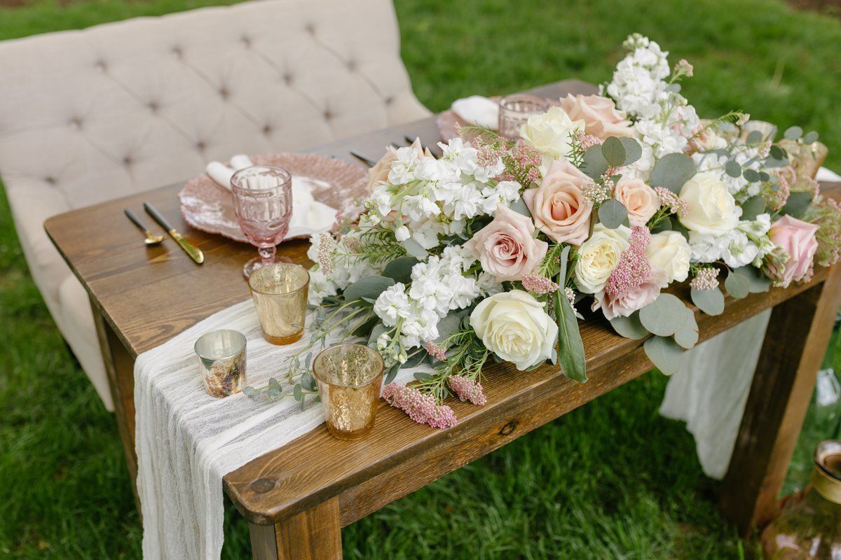 Outdoor rustic table with blush and white floral centerpiece, candles, and place settings beside a tufted bench
