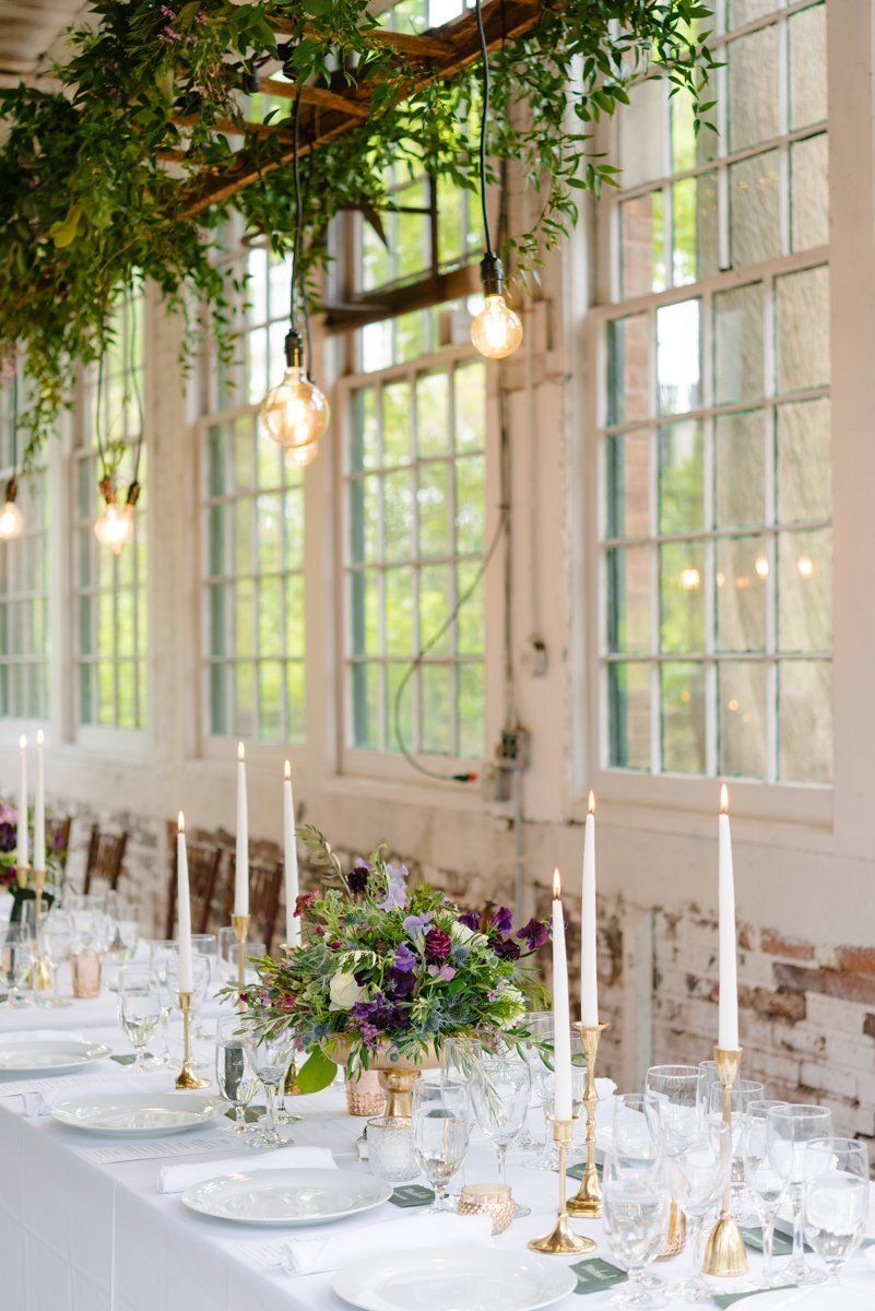 Elegant long table set with white plates, candles, and flowers in a bright room with hanging greenery.