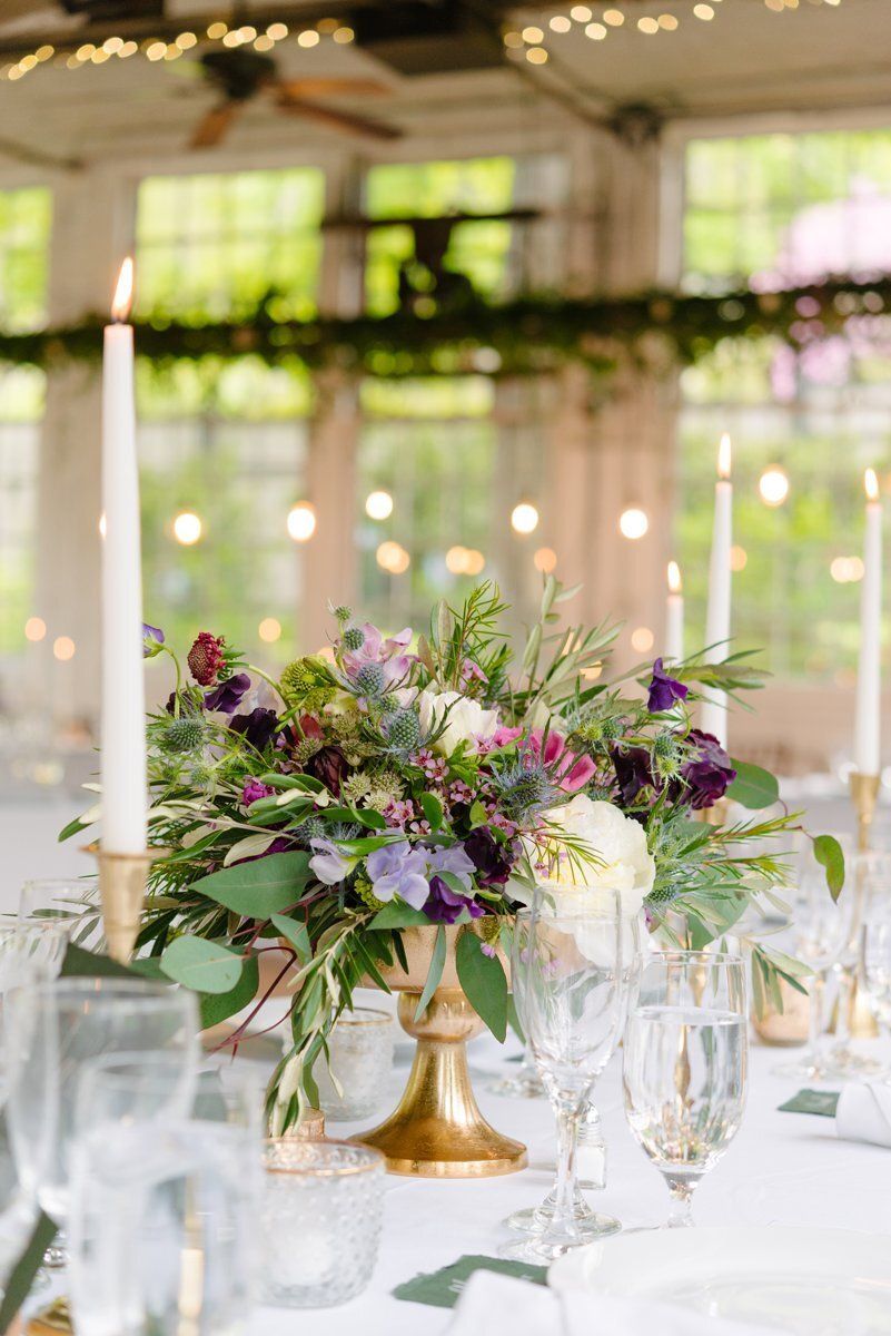 Elegant wedding reception table with a gold vase of purple and white flowers, candles, and glassware