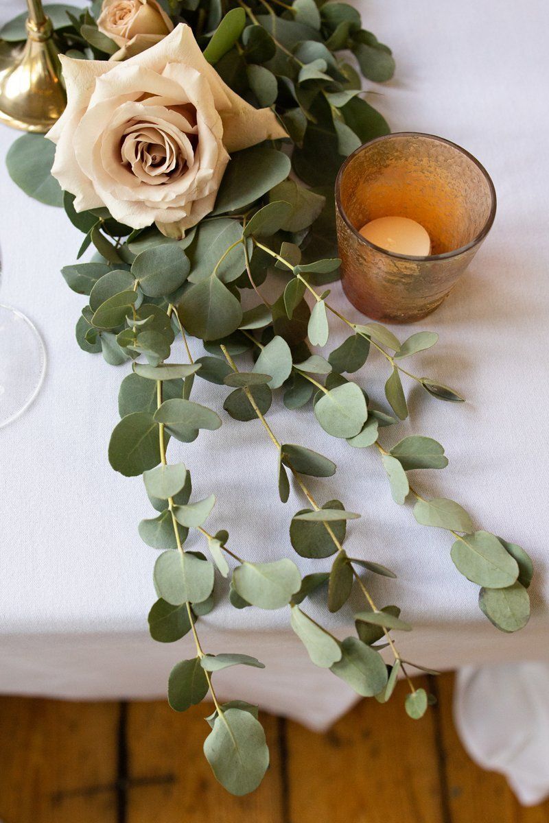 Beige rose and eucalyptus greenery beside a lit candle on a white tablecloth