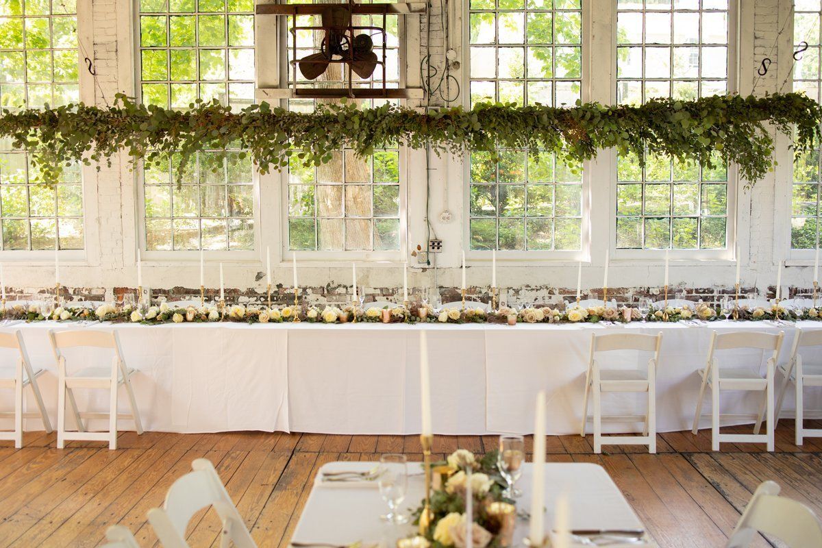 Bright wedding reception table with white chairs, greenery garlands, and large windows in a sunlit hall