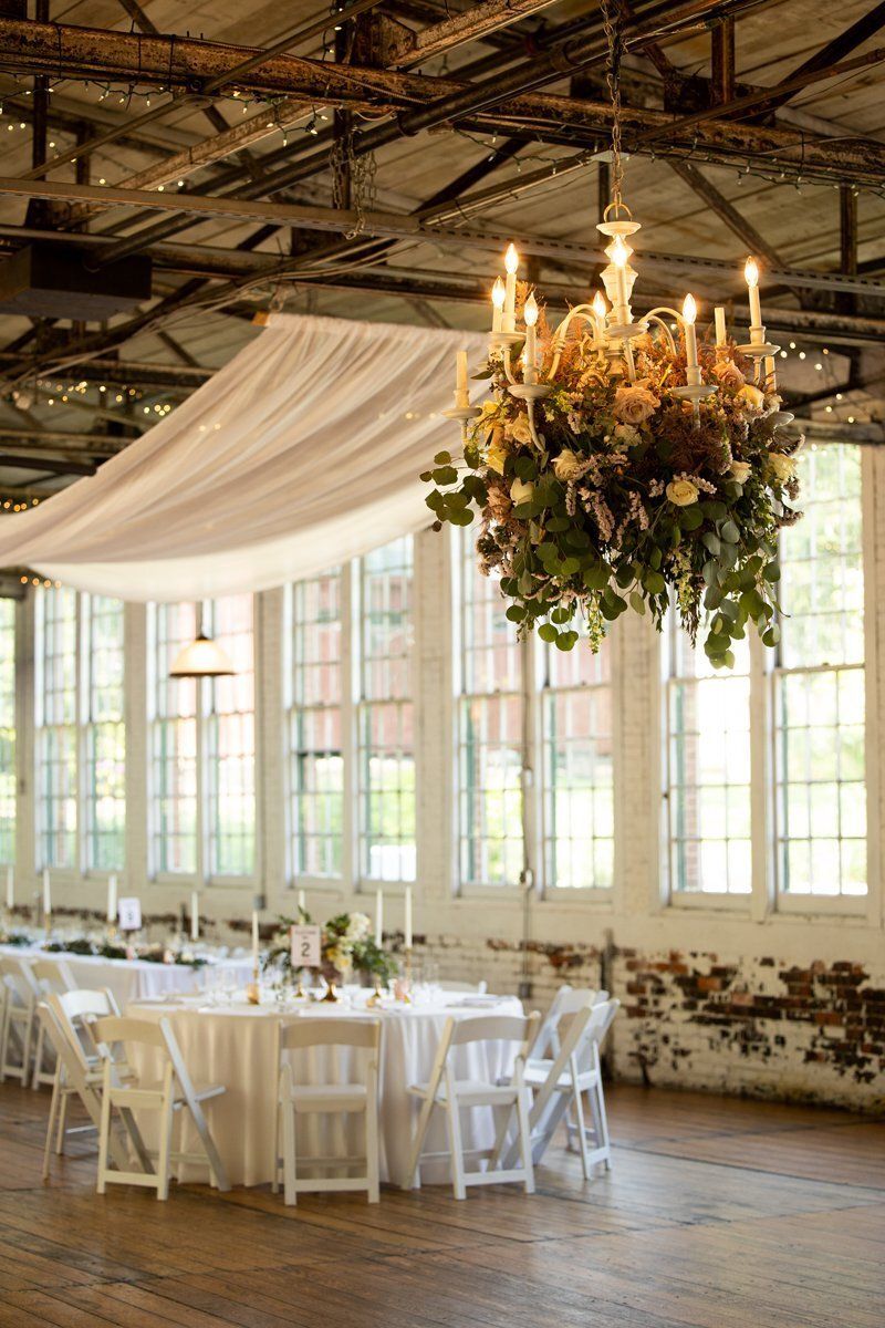 Elegant banquet setup in a rustic hall with white tables, draped fabric, and a chandelier centerpiece