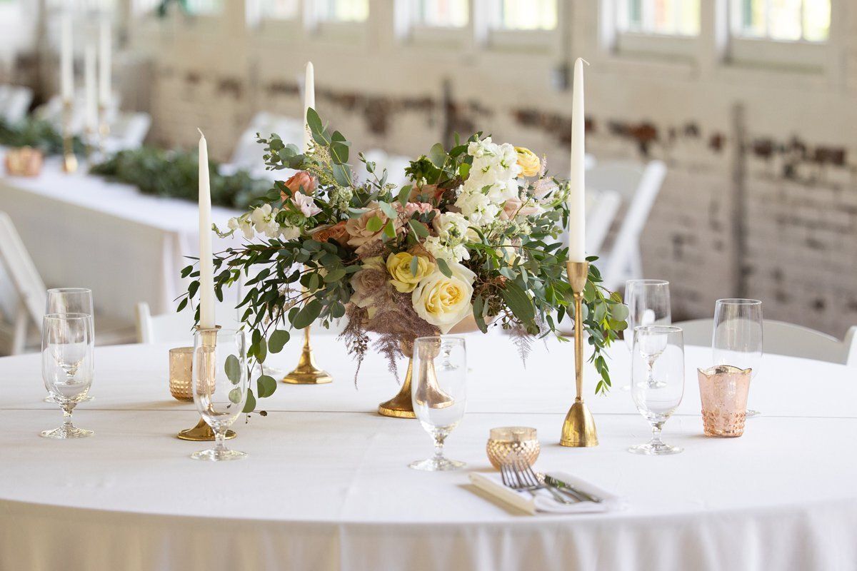 Elegant banquet table with white linens, floral centerpiece, candles, and glassware in a bright reception hall