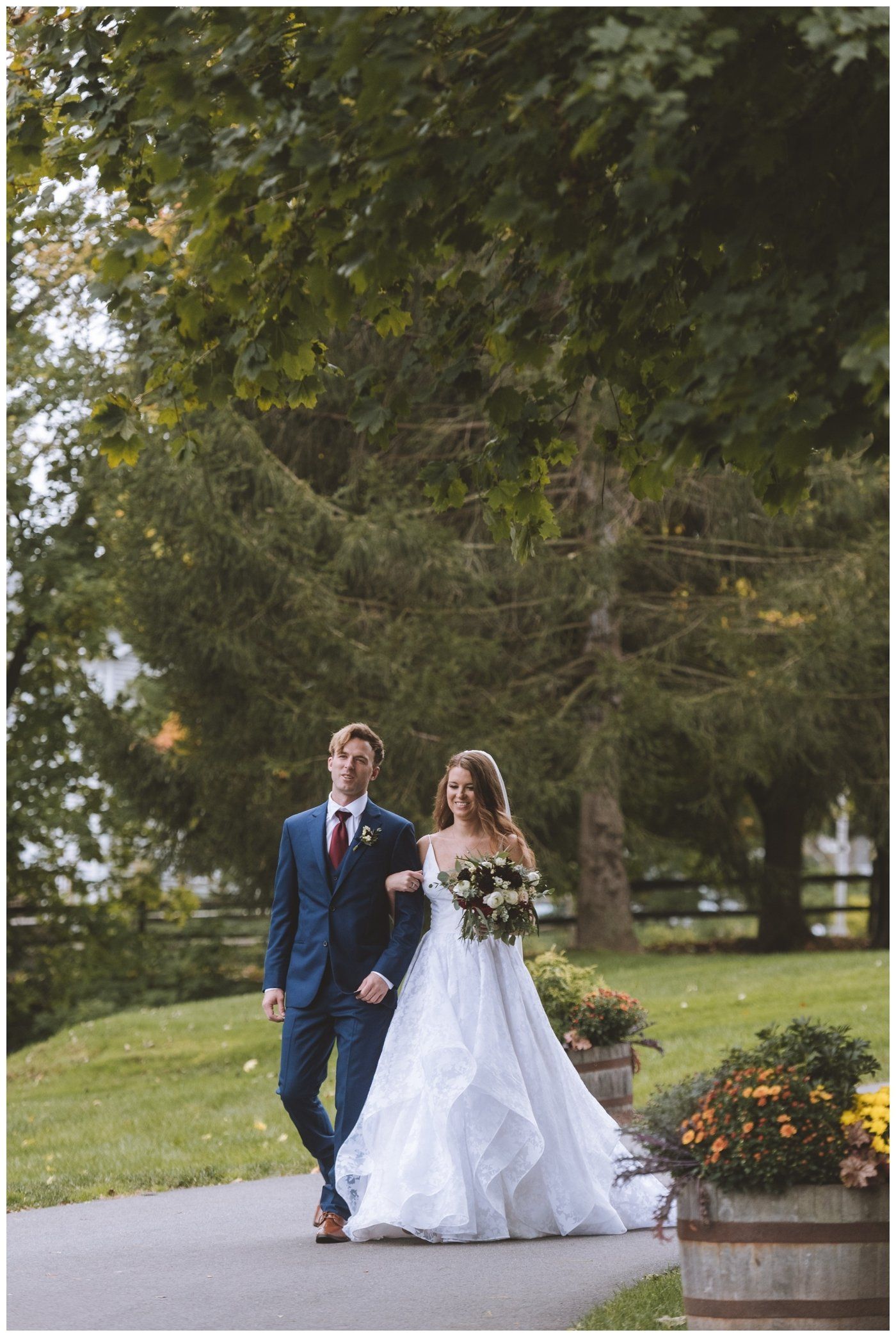 Bride and groom walking on a path in a garden, bride in white gown holding bouquet, groom in blue suit