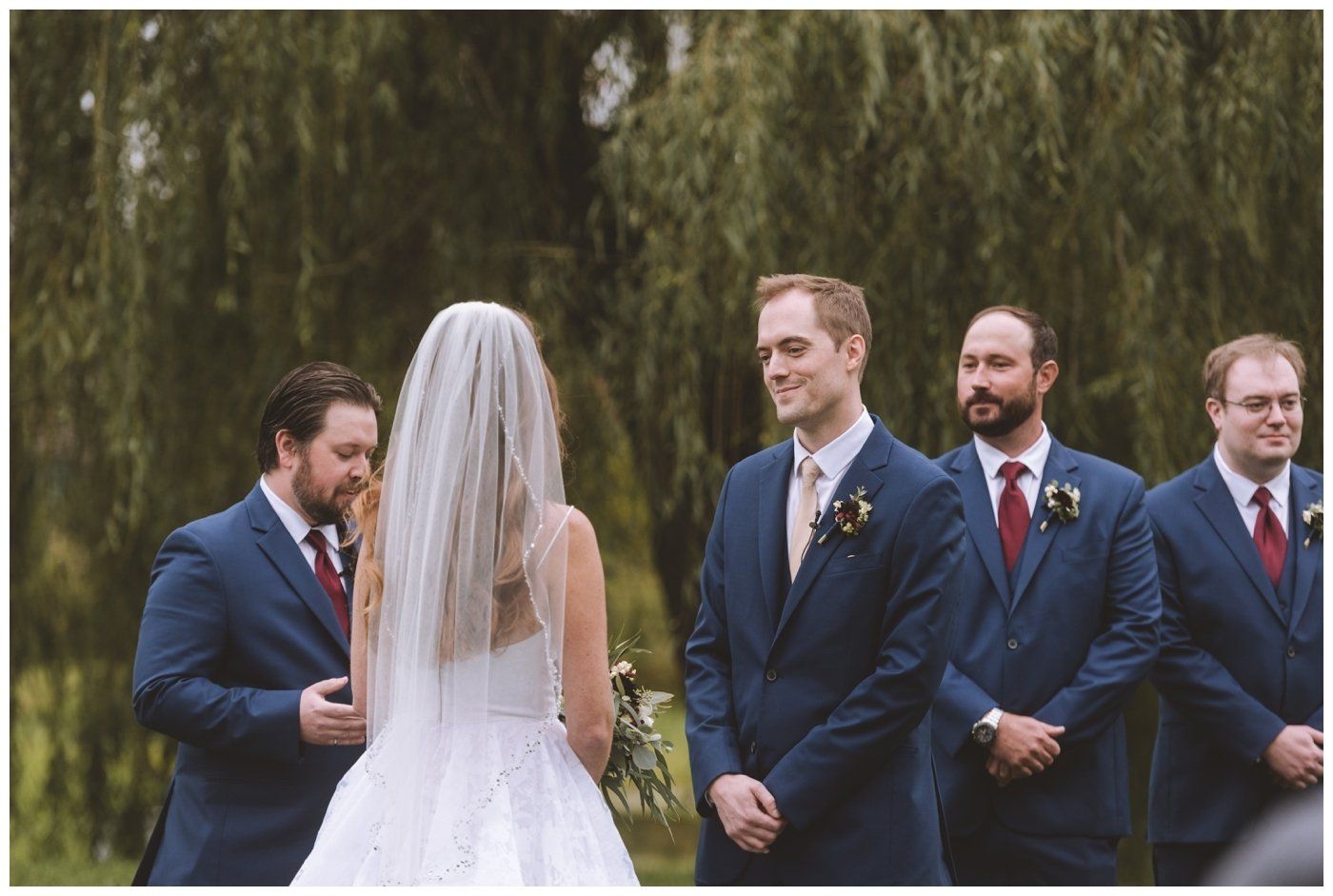 Wedding party stands outdoors under trees; bride faces groom in blue suit, attendants nearby.