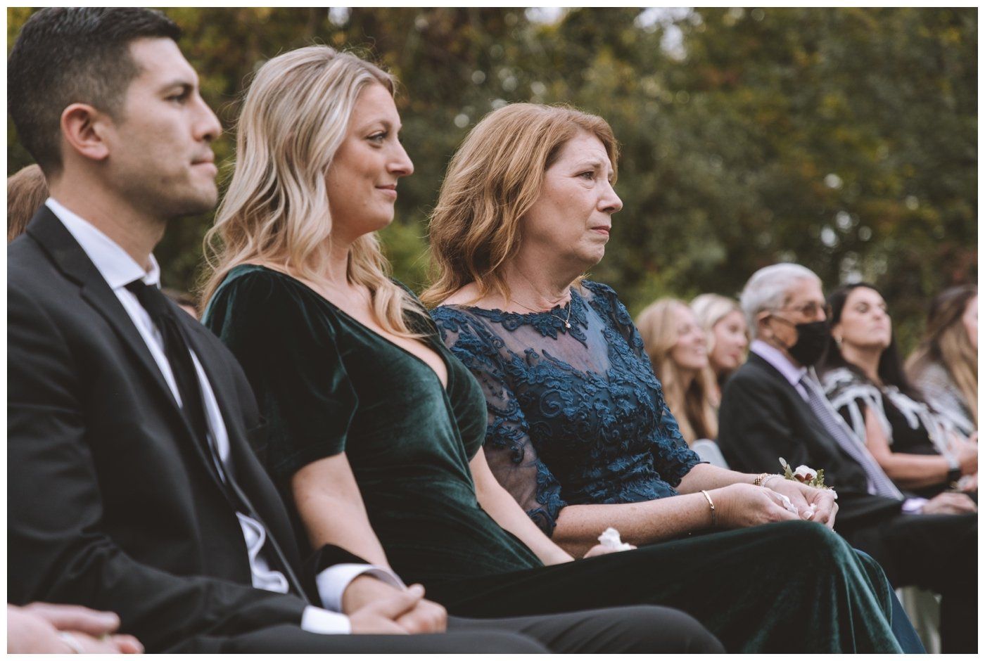 People seated in formal attire at an outdoor ceremony, watching attentively