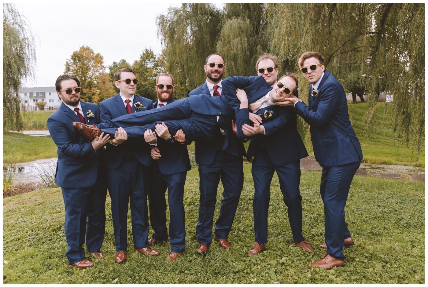 Six men in blue suits and sunglasses posing playfully outdoors, holding one man horizontally.