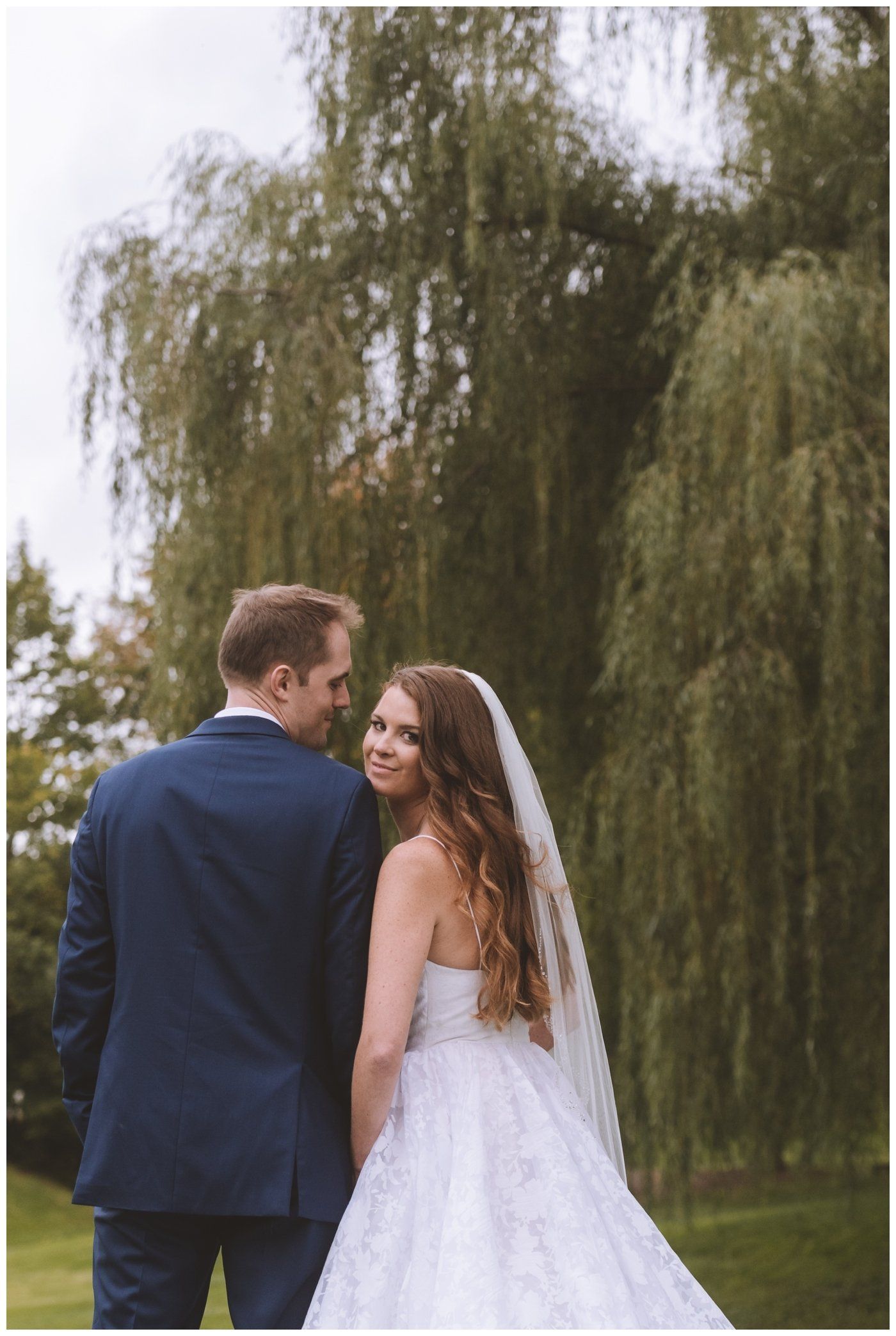 Bride and groom looking back over their shoulders in a garden with a weeping willow tree