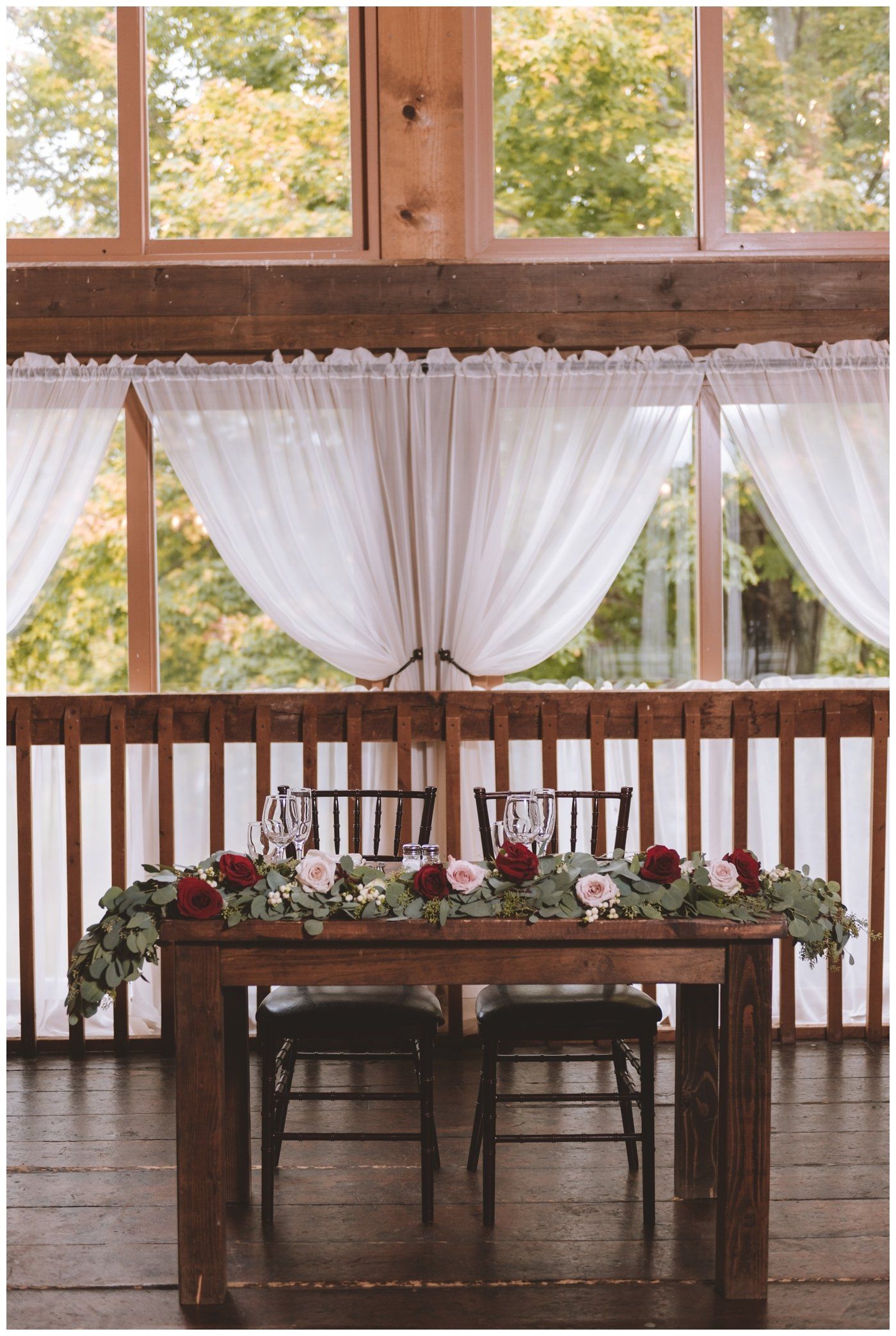 Rustic indoor wedding reception table with flowers, white drapes, and wooden chairs by large windows
