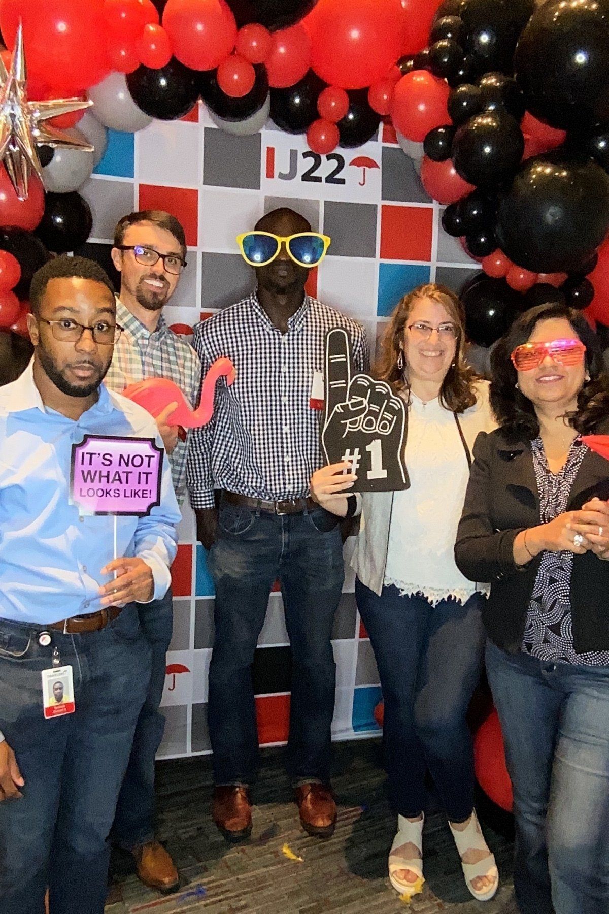 Five people posing at a party booth with black and red balloons, holding signs and a trophy.