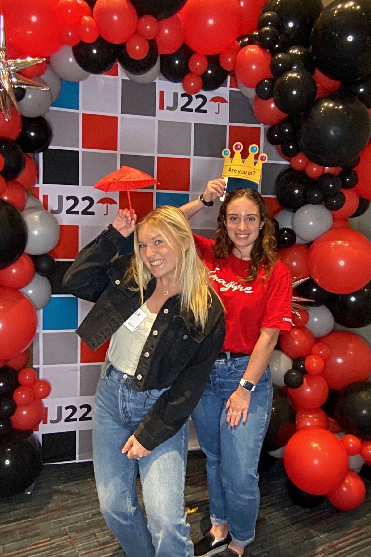 Two people posing in front of a red and black balloon arch and J22 backdrop, smiling at a party