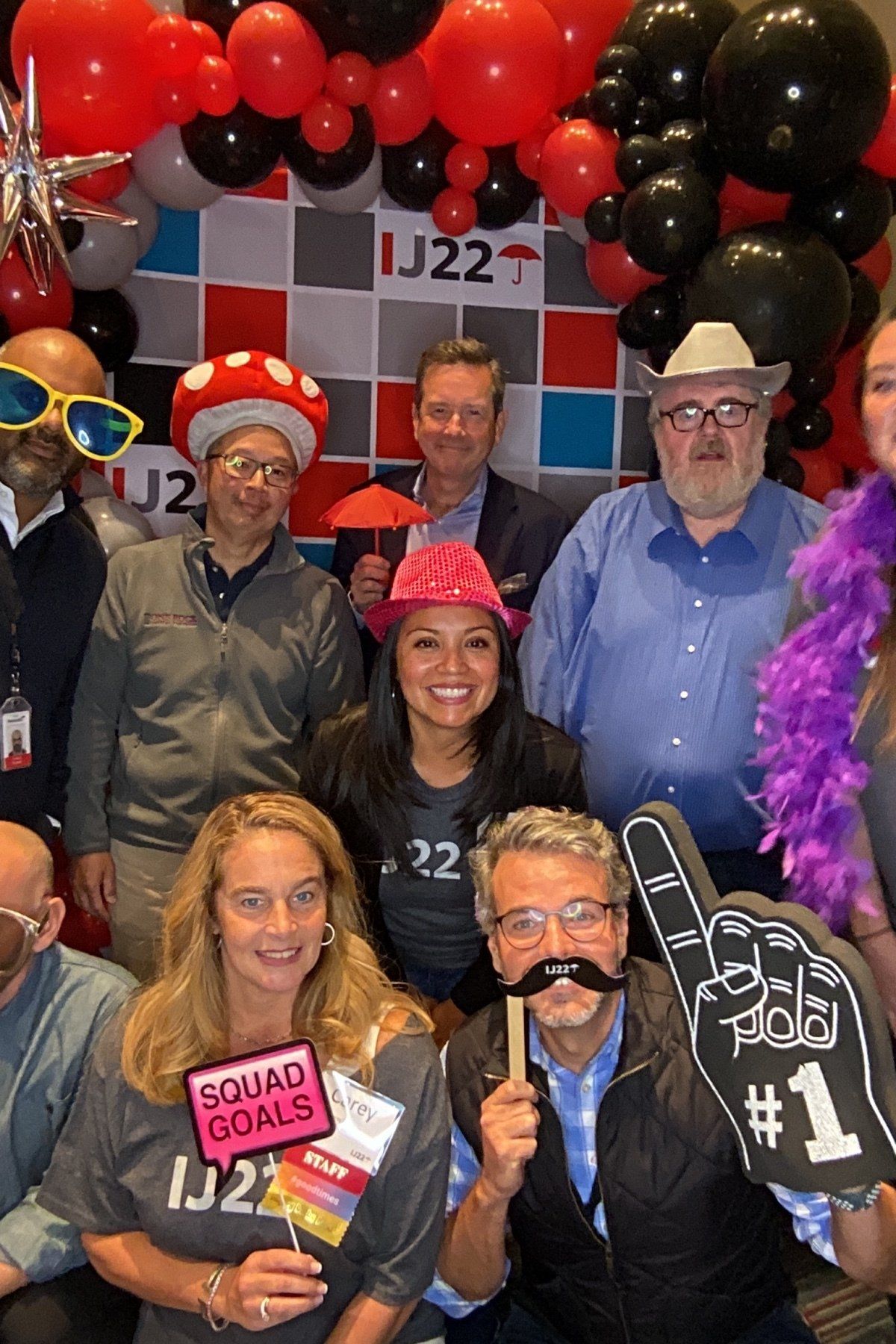 Group posing at a festive event with balloons, hats, and playful props in front of a J22 backdrop