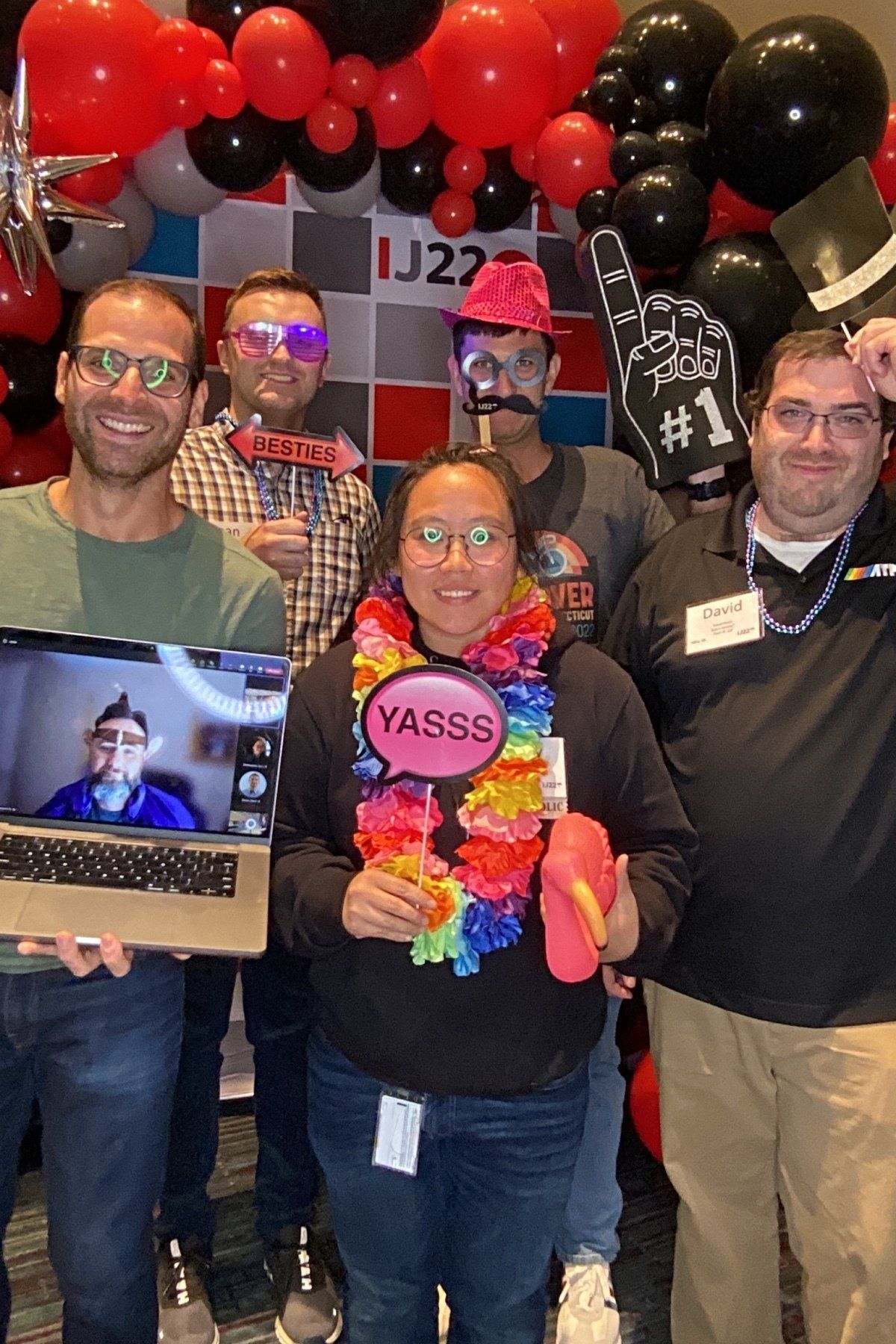 Group of smiling people at an event, holding balloons and a colorful YASSS sign, with a small screen in front.