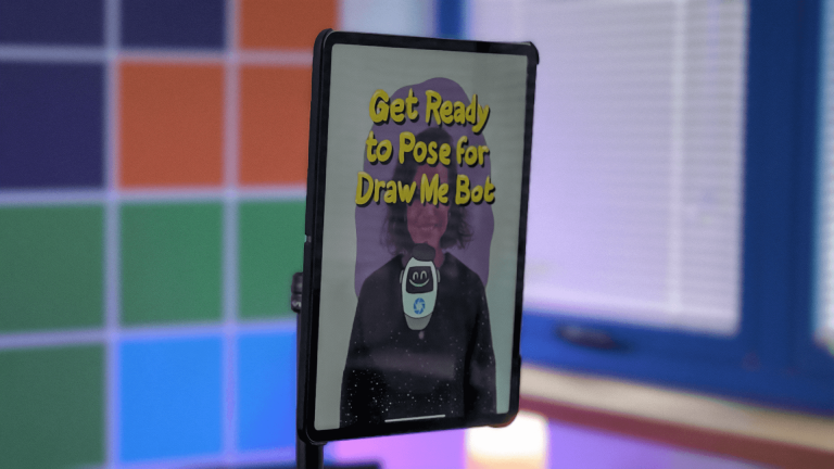Phone on stand in front of colorful grid background, screen showing “Get Ready to Pose for Draw Me Bot”
