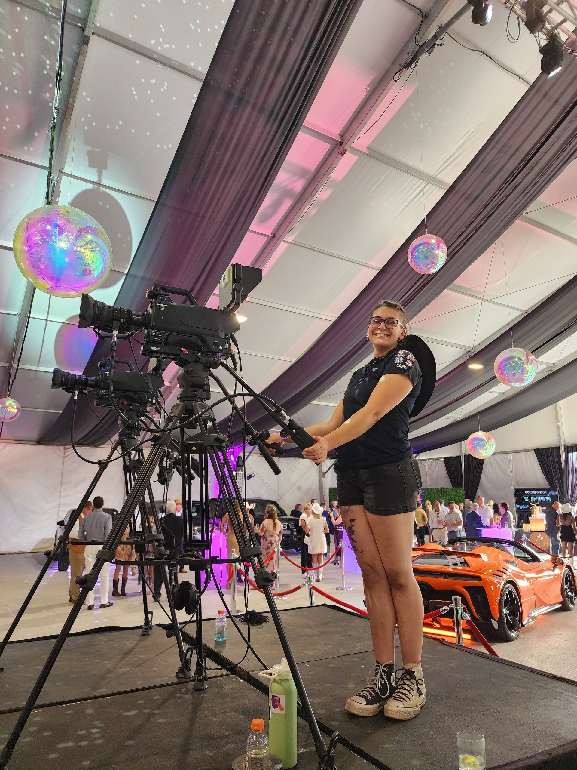 Person standing by a large camera rig at an indoor event, with colorful hanging lights overhead