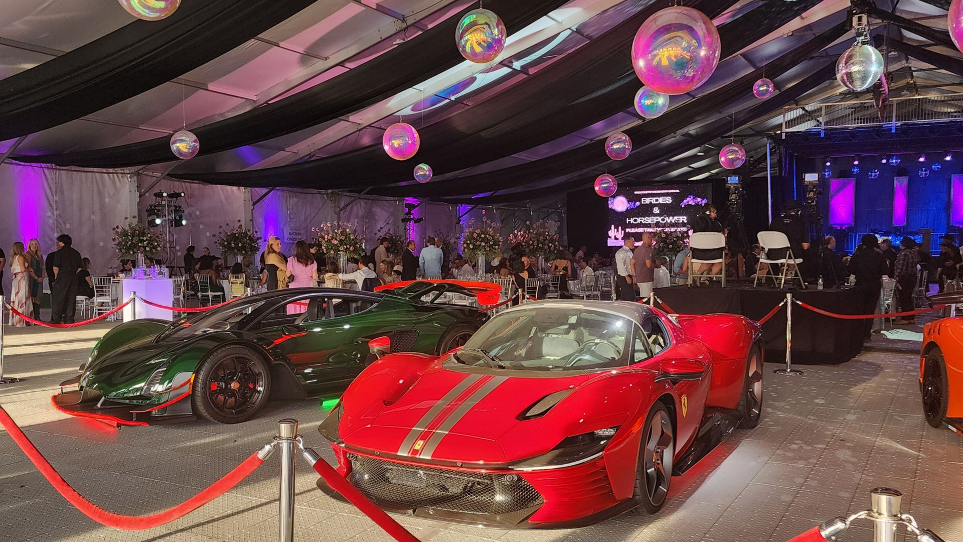 Two red sports cars displayed under purple lights at an indoor event with velvet ropes