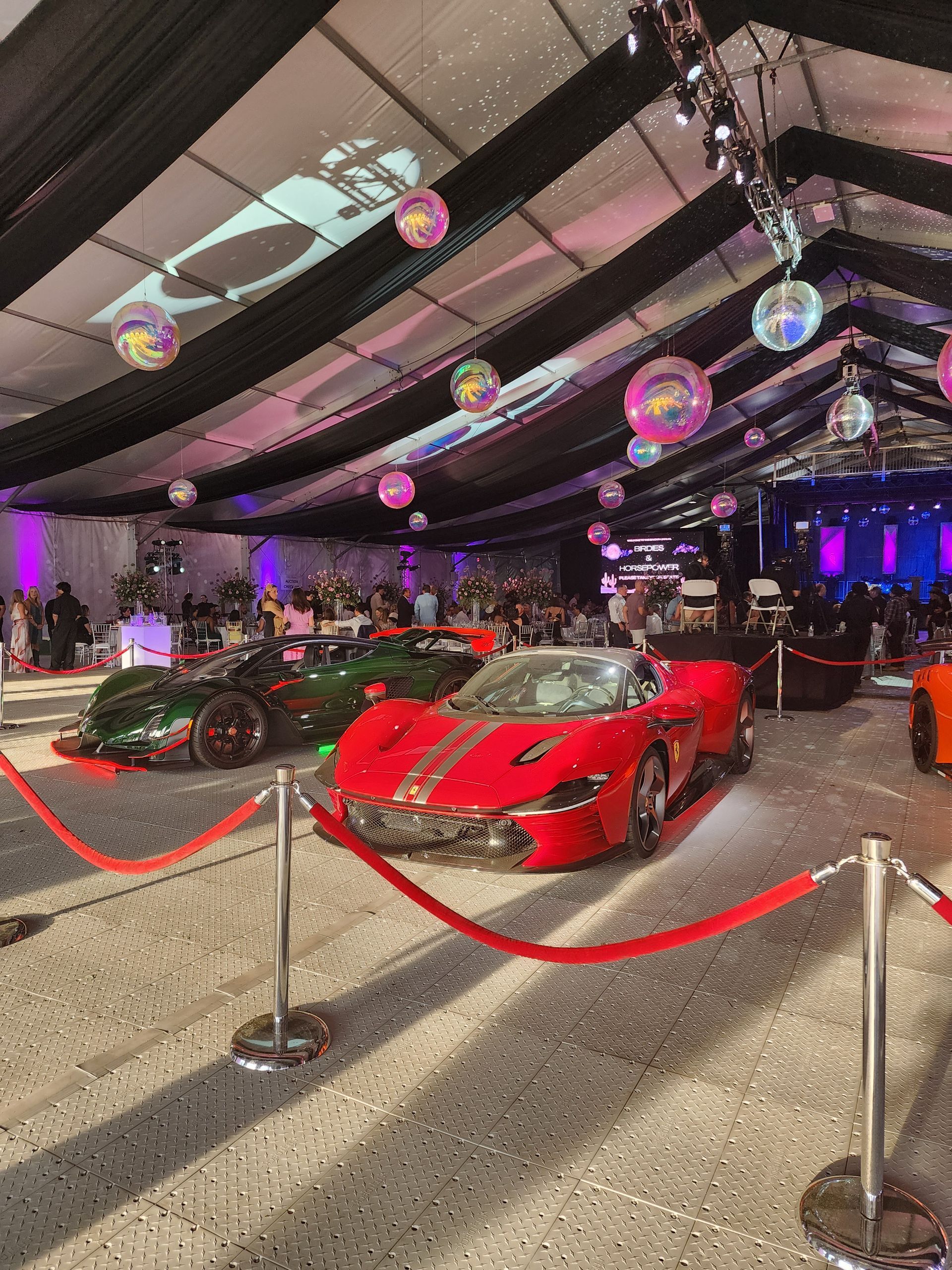 Red sports car on display under a striped tent with hanging lights and event barriers