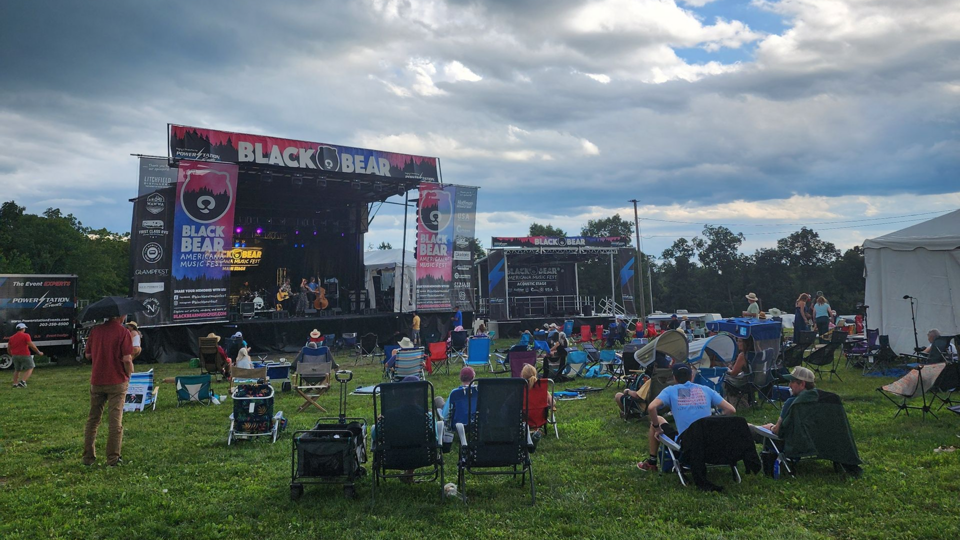 Outdoor music festival crowd seated on grass facing a large stage under cloudy skies