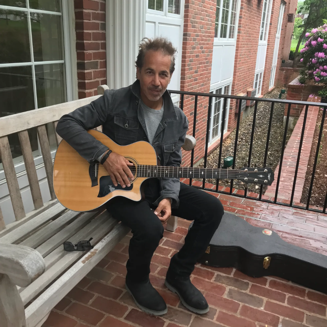 Man sitting on a porch bench holding an acoustic guitar beside an open guitar case.