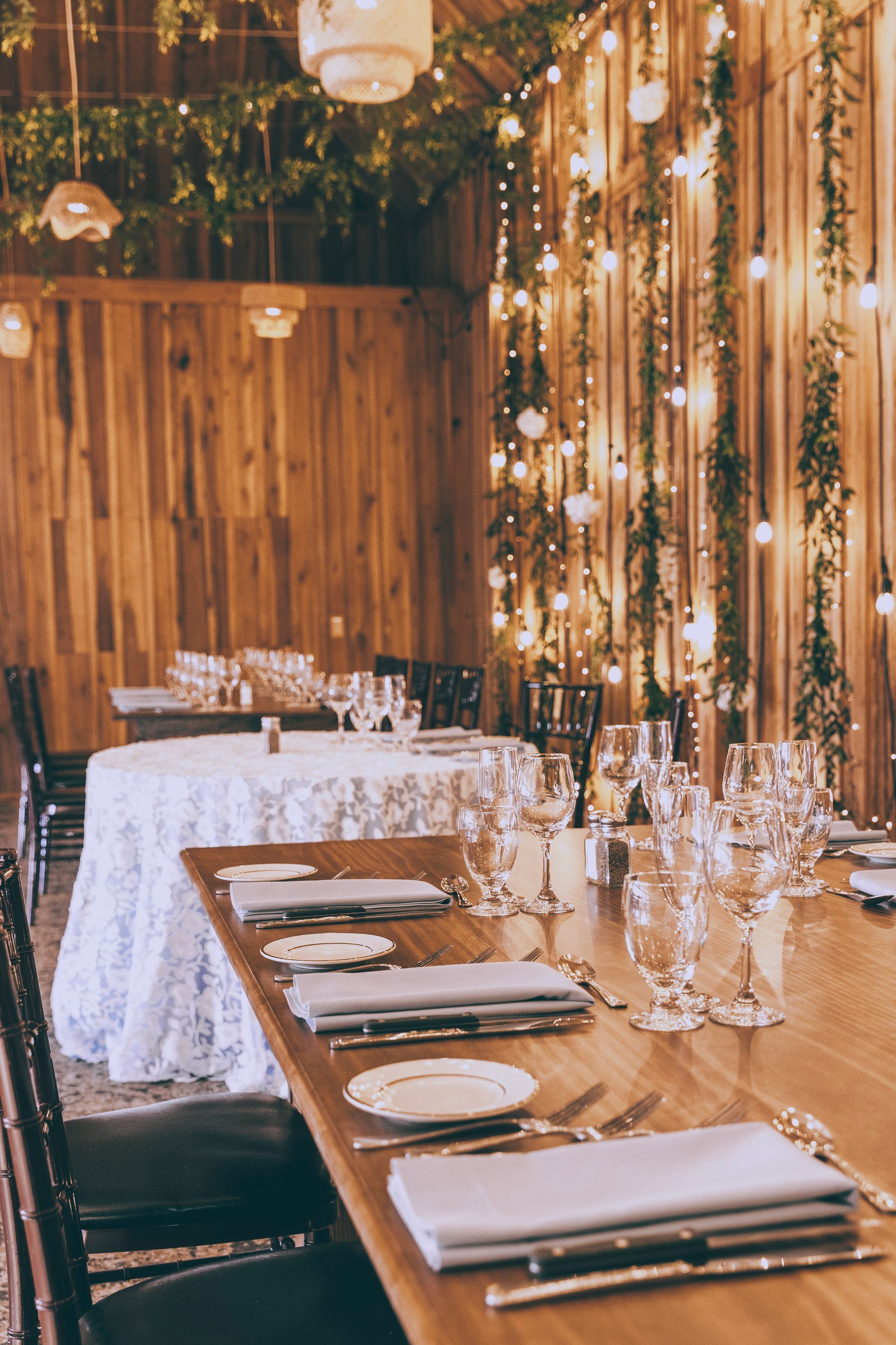 Rustic banquet table set with white plates, glassware, and candles under warm string lights