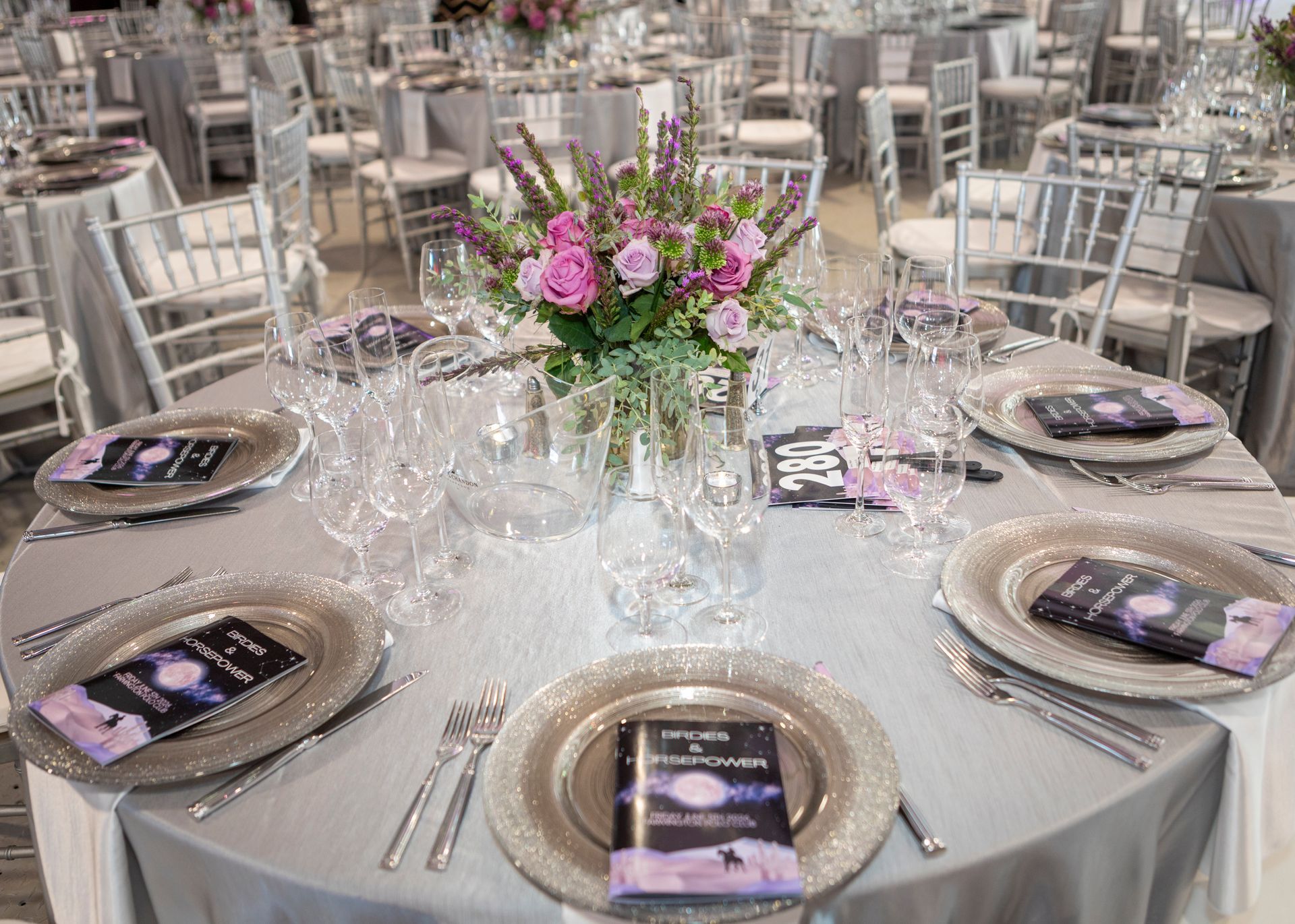 Banquet table set with silver chargers, glasses, and pink floral centerpiece in a bright reception hall