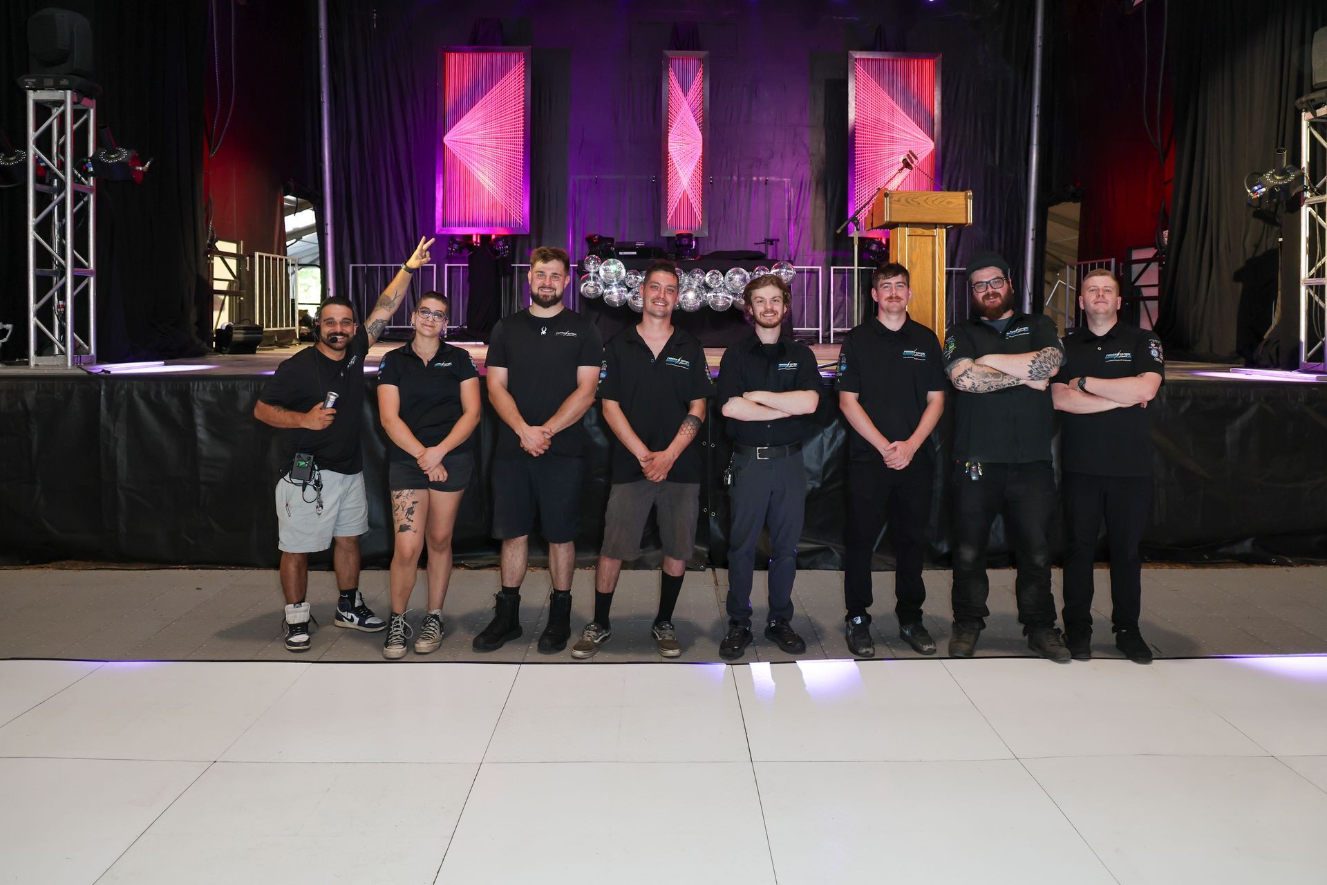 Group of people standing on a stage in black shirts, with colorful lit panels and curtains behind them.