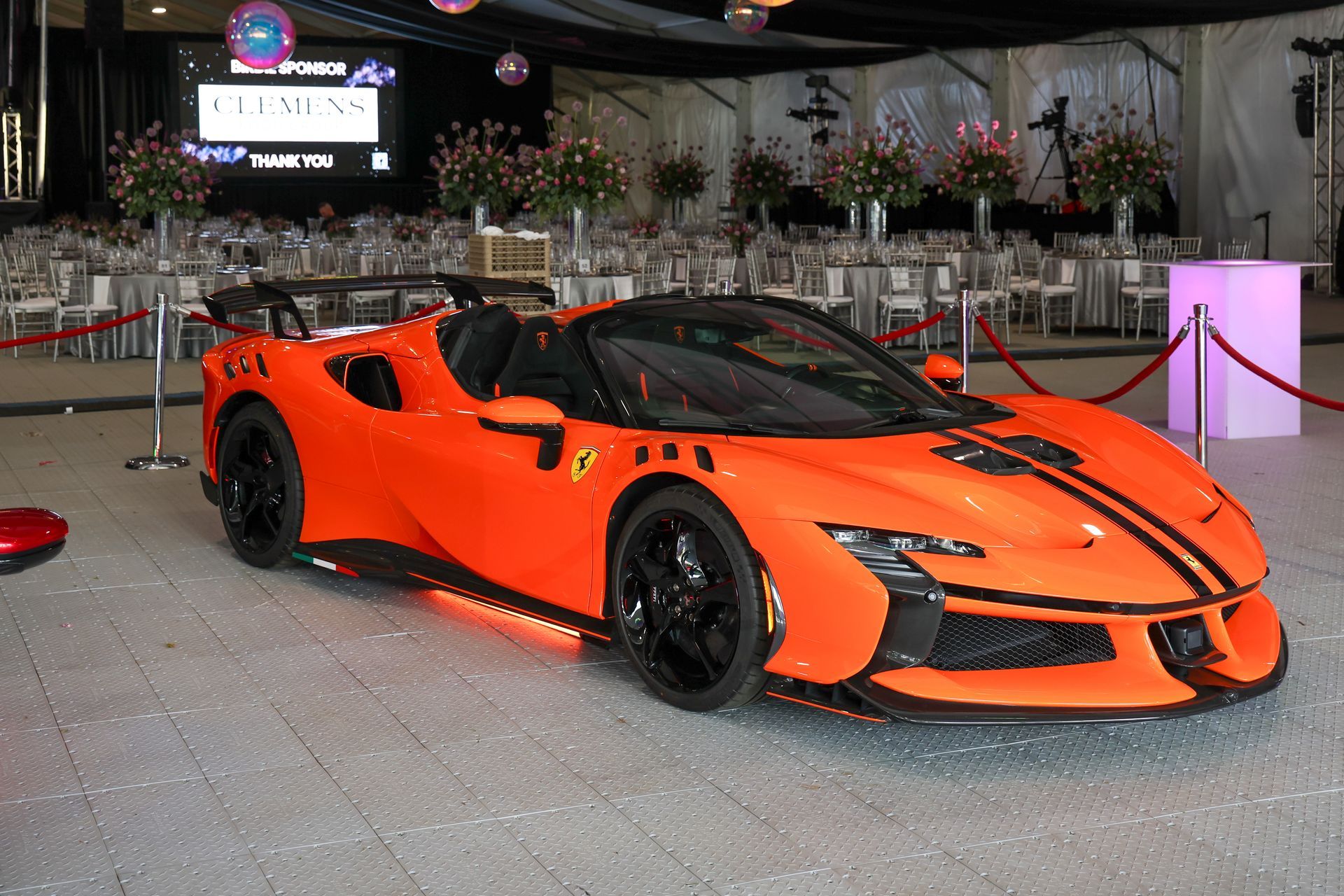 Orange sports car on display indoors at an event, with black racing stripes and a glossy floor.
