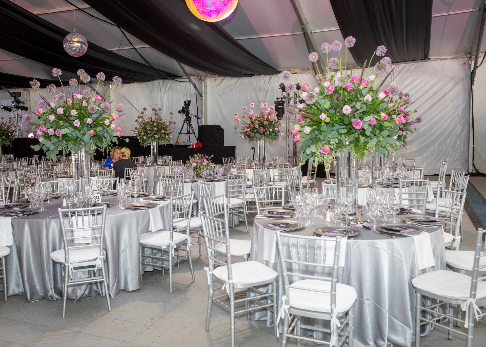 Elegant outdoor reception with round tables, white chairs, and tall pink floral centerpieces under a tent.