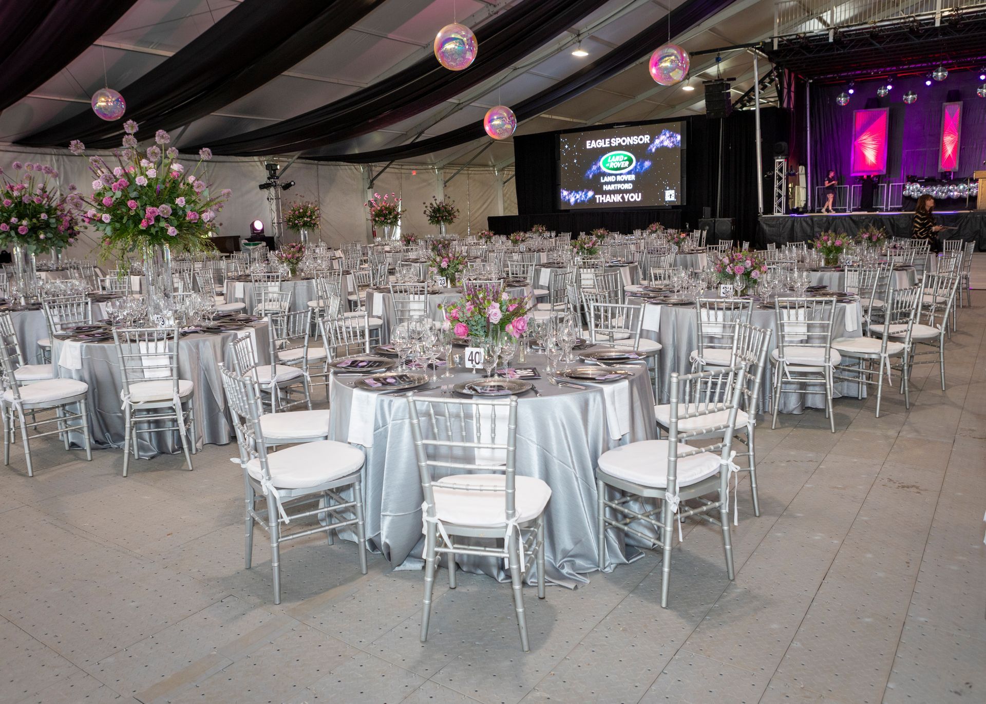 Banquet hall with round tables, silver chairs, floral centerpieces, and a stage with purple lighting