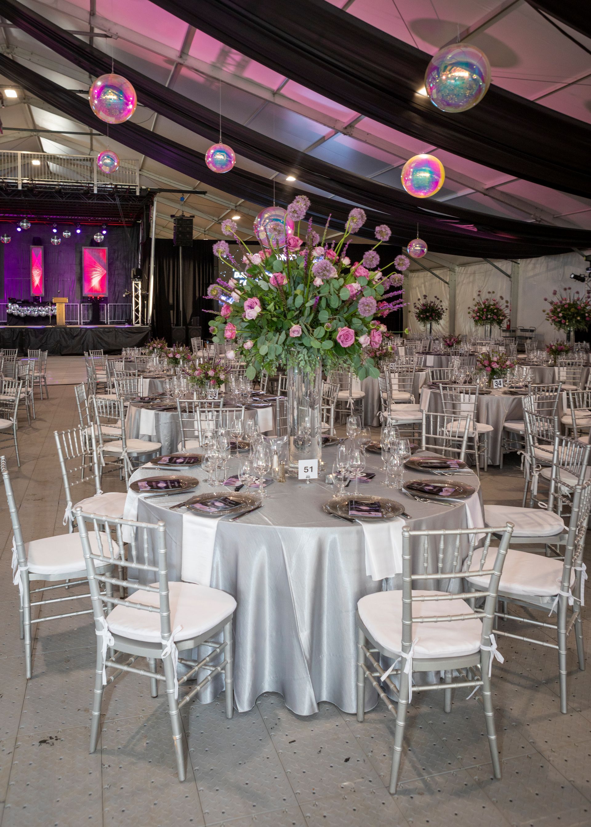 Elegant banquet hall with round tables, silver chairs, and pink floral centerpieces under draped ceiling lights