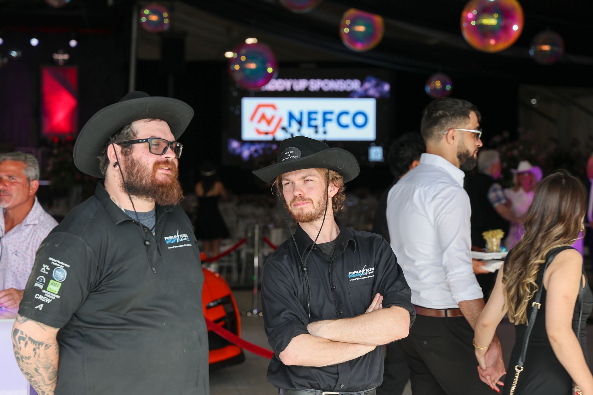 People chatting at a dimly lit event, with a sponsor sign and bubbles in the background