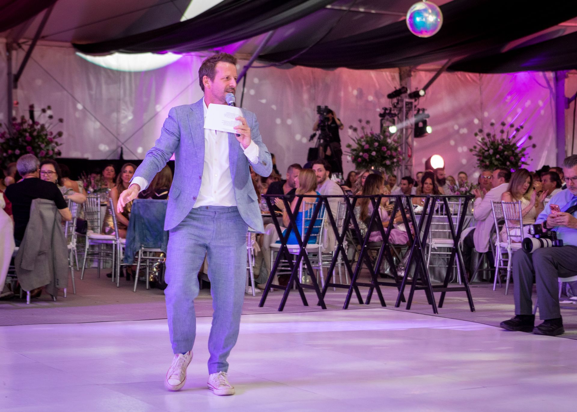 Man in a light blue suit singing on a dance floor at a purple-lit wedding reception