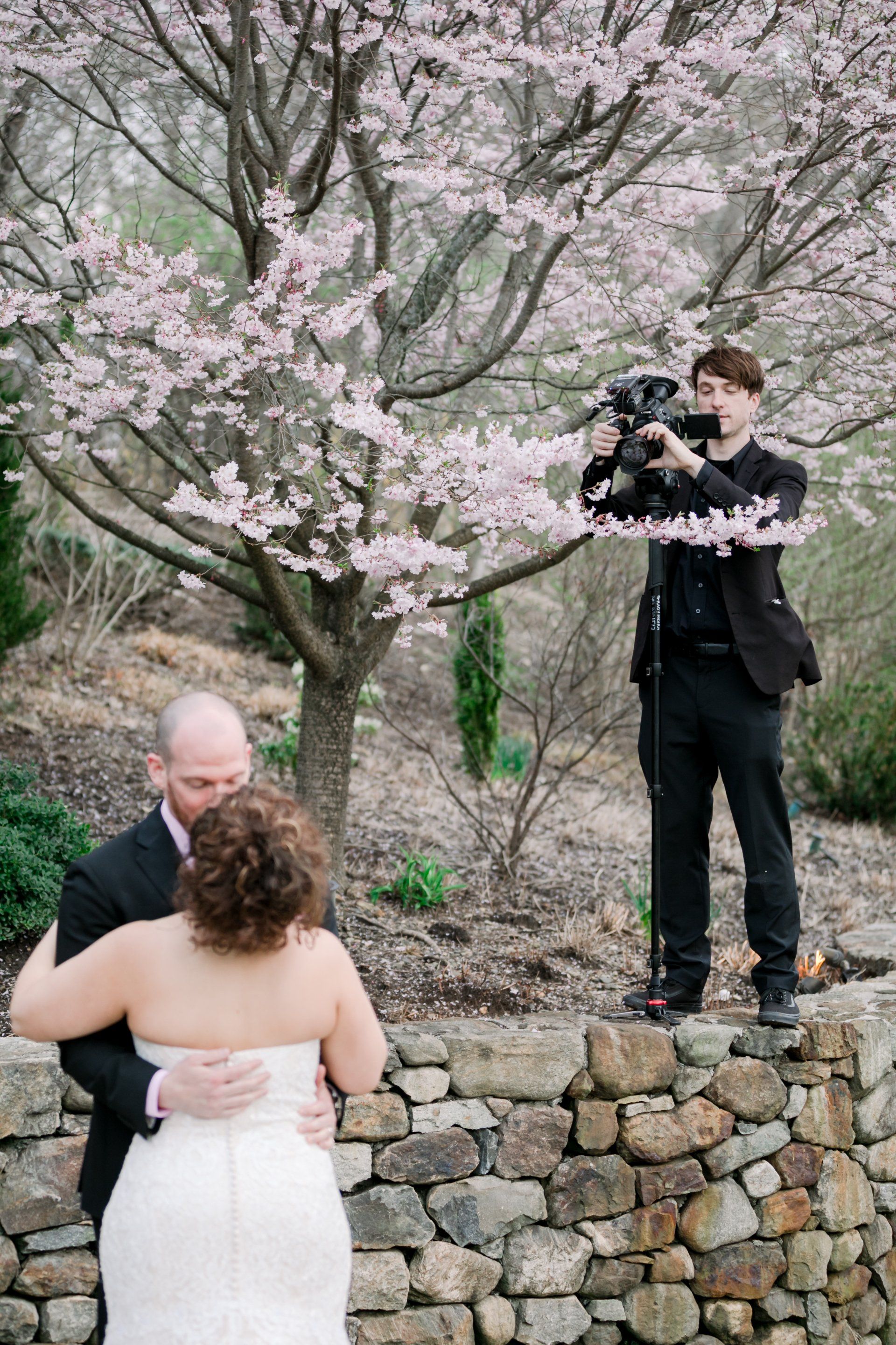 Couple exchanging vows outdoors while a photographer captures the moment by a cherry blossom tree