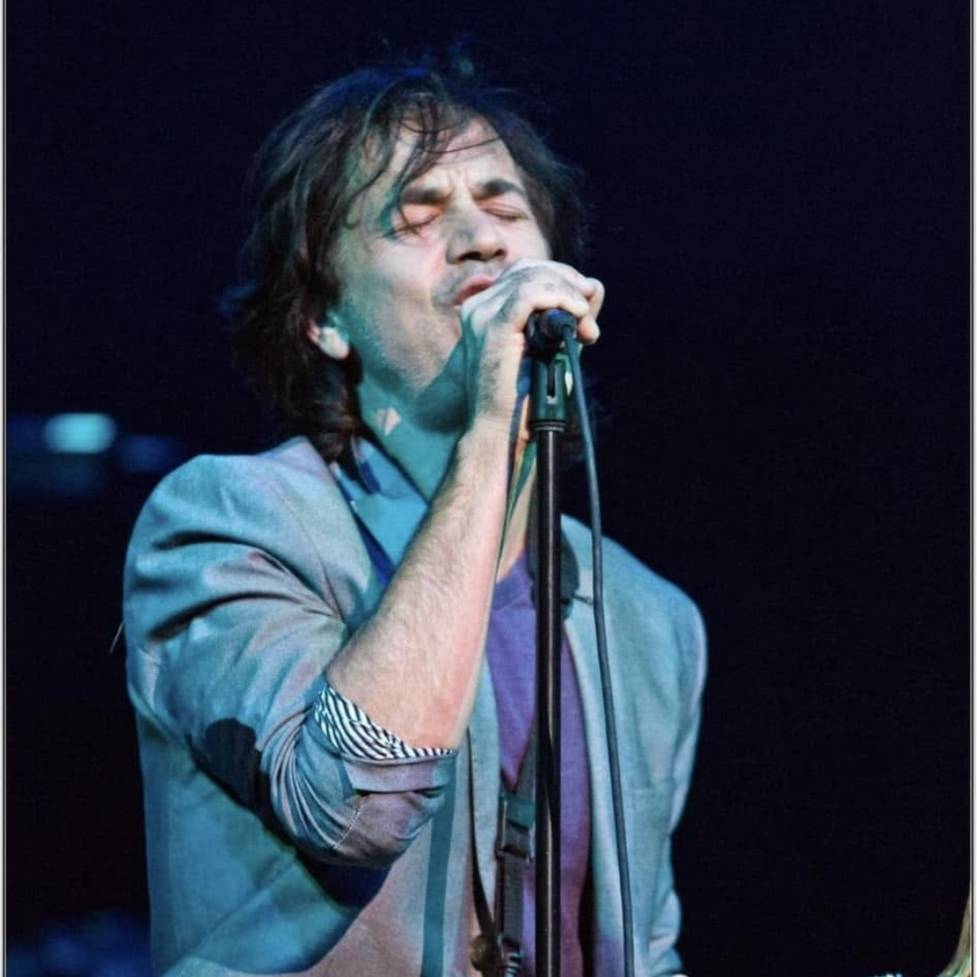 Singer performing into a microphone on stage under blue lighting, eyes closed and wearing a gray jacket